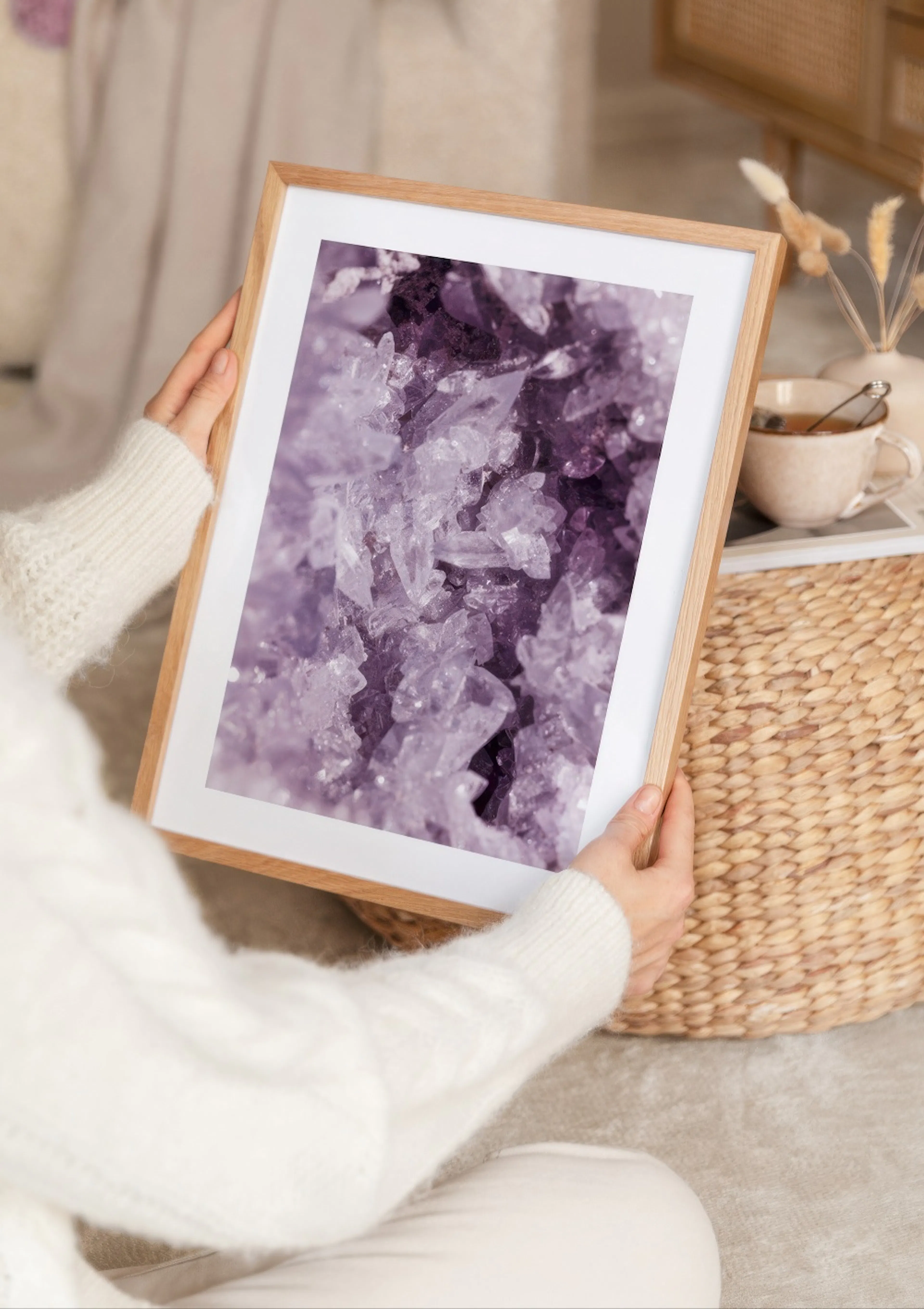 A person in a white jumper holds a poster of purple amethyst crystals, with a woven basket and teacup in the background.