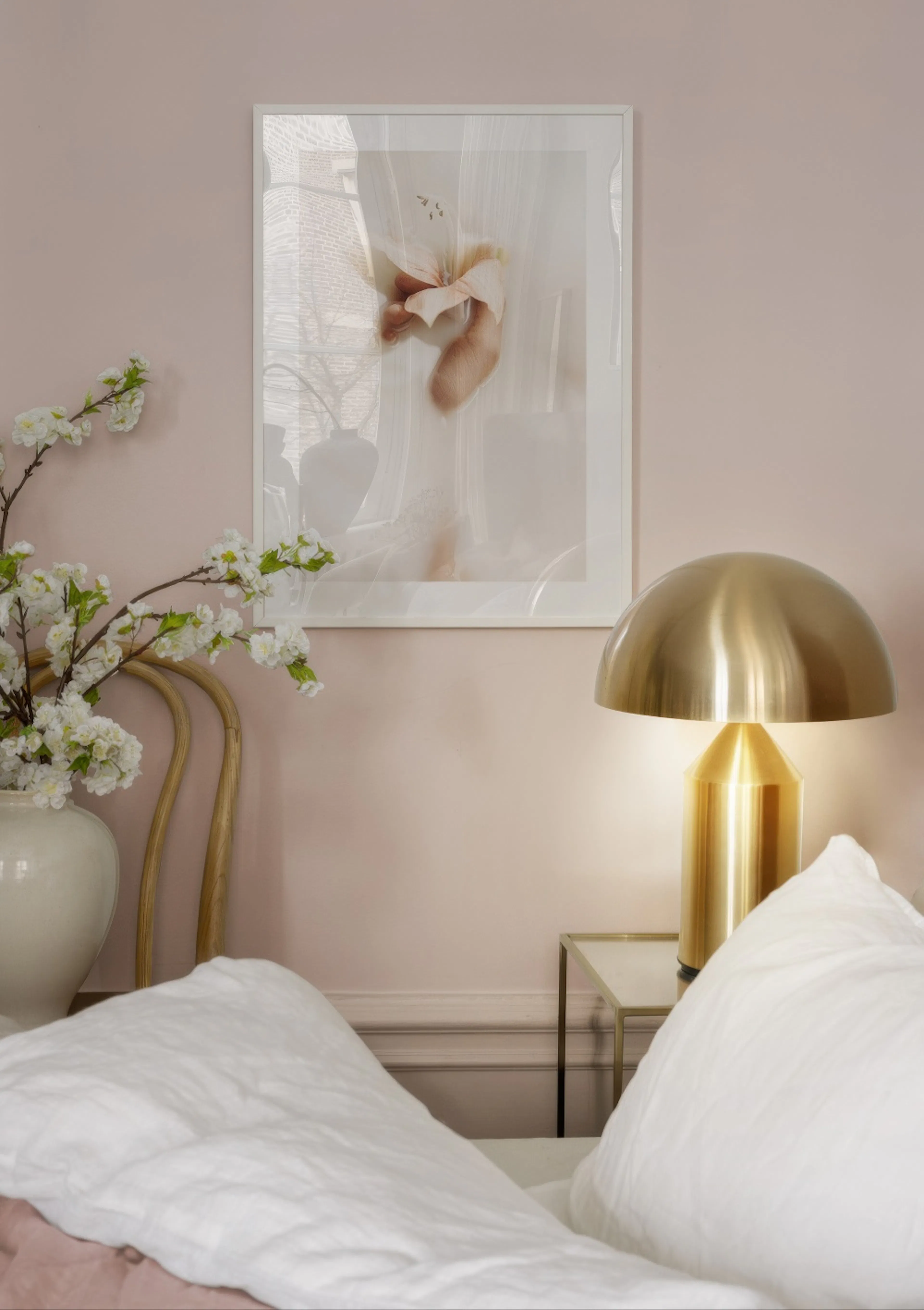 연한 분홍색 벽에 걸린 은은한 연분홍색 꽃 포스터. 침실의 침대와 황금색 스탠드 옆에 놓여있다.
