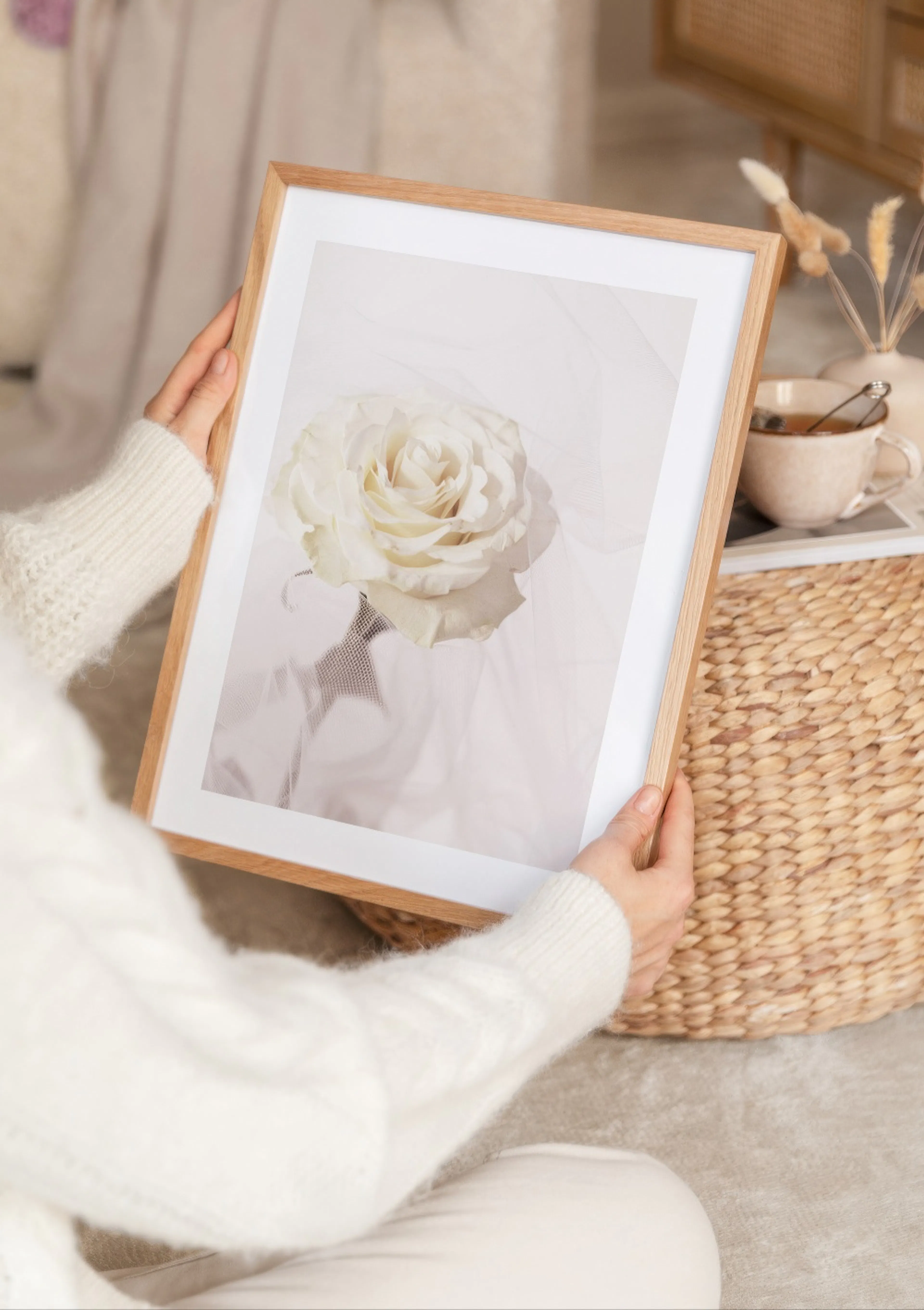 A person in a white jumper holds a poster of a delicate white rose, with a woven basket and cup of tea nearby.