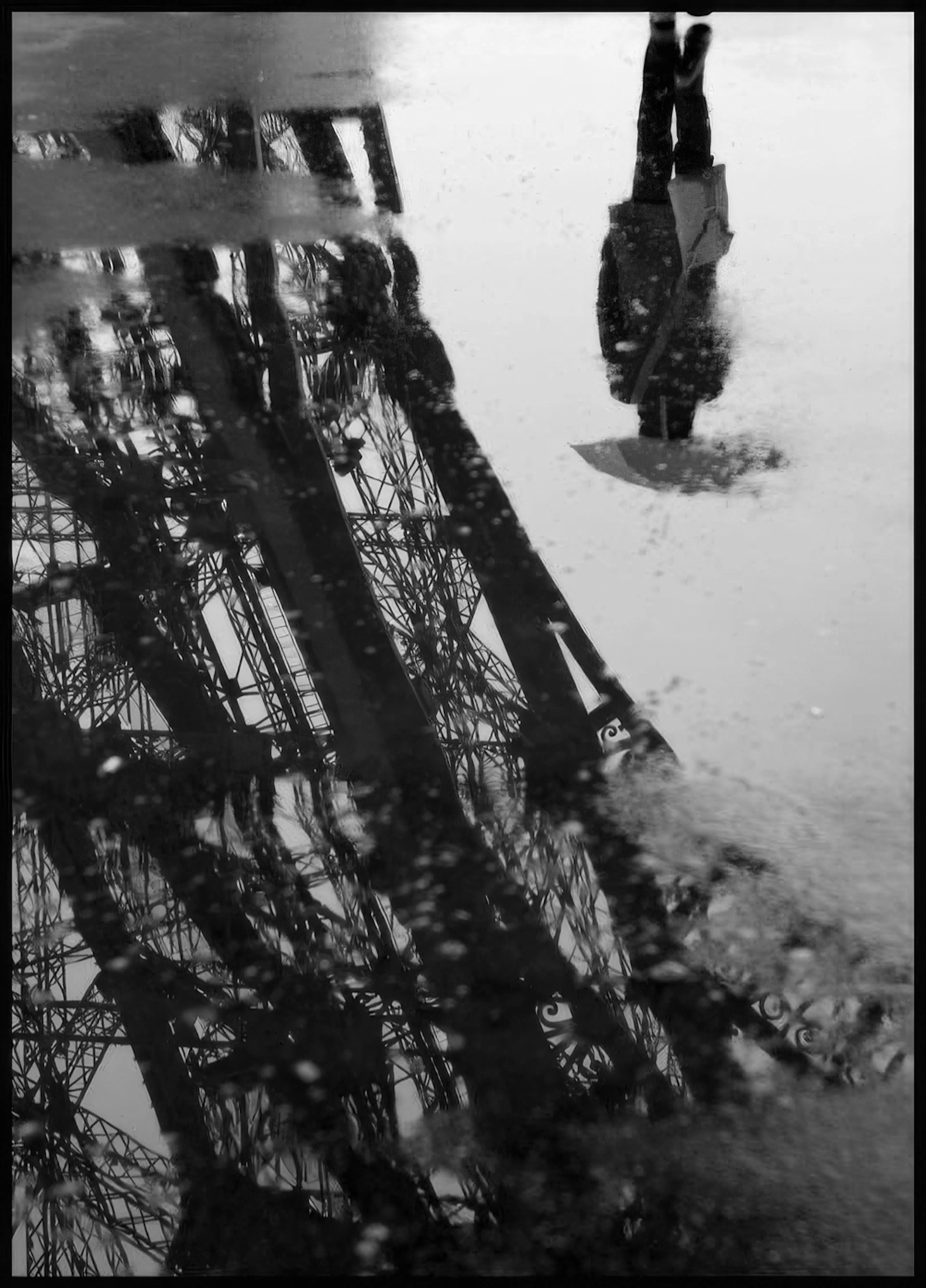 Mustavalkoinen juliste, jossa Eiffel-torni heijastuu lätäköstä ja sateen kastelema jalankulkija näkyy ylhäällä.