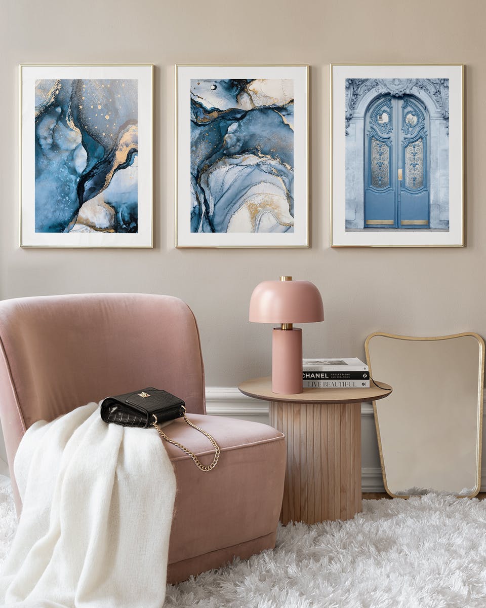 Három poszter egy szobában. Két absztrakt kék-arany festmény és egy kék ajtó poszter egy rózsaszín fotel fölött.