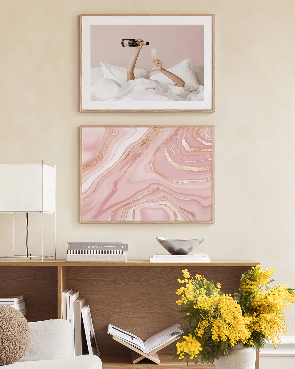 A gallery wall with two posters: a pink marble poster with gold veins, and a poster of hands pouring champagne in bed, displayed