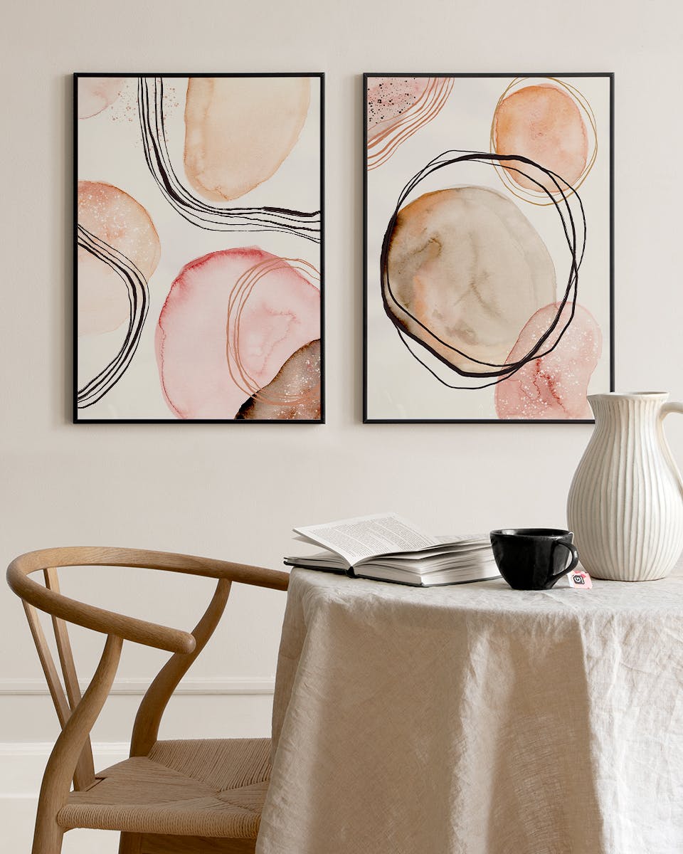 Due pacchetti di poster con forme astratte acquerellate in rosa e beige, linee nere e oro, appesi sopra un tavolo con libro e ta