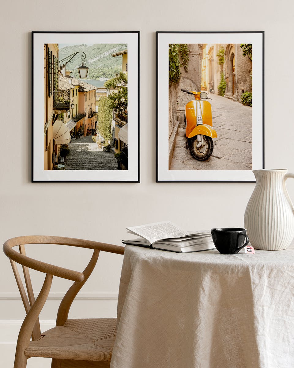 Zwei gerahmte Poster einer italienischen Gasse und eines Rollers, über einem Tisch mit Buch und Krug.