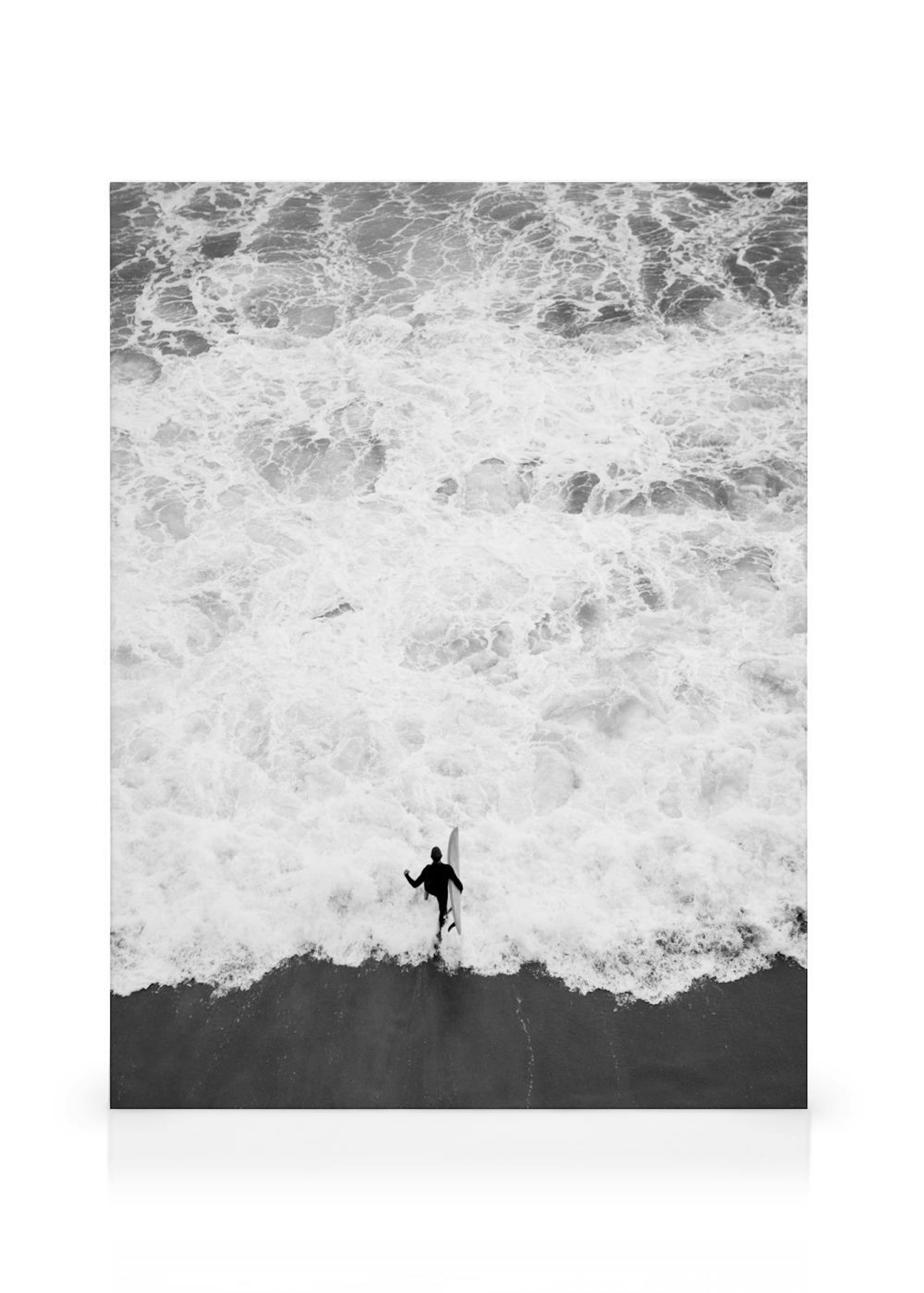 Black and white print of a lone surfer walking into crashing ocean waves from a dark sandy beach.