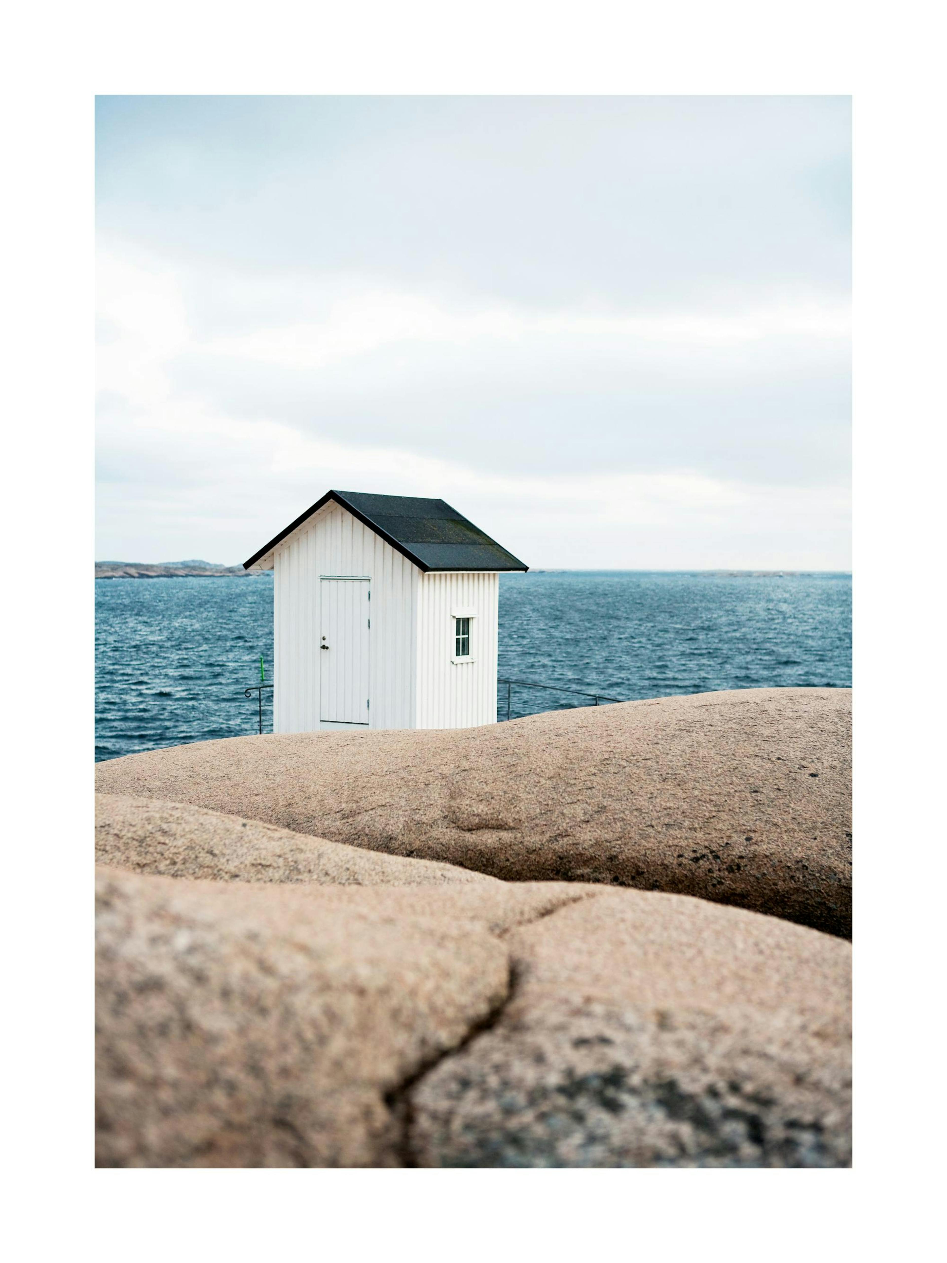 白い小屋と青い海、手前に岩が写る美しい風景のポスター。