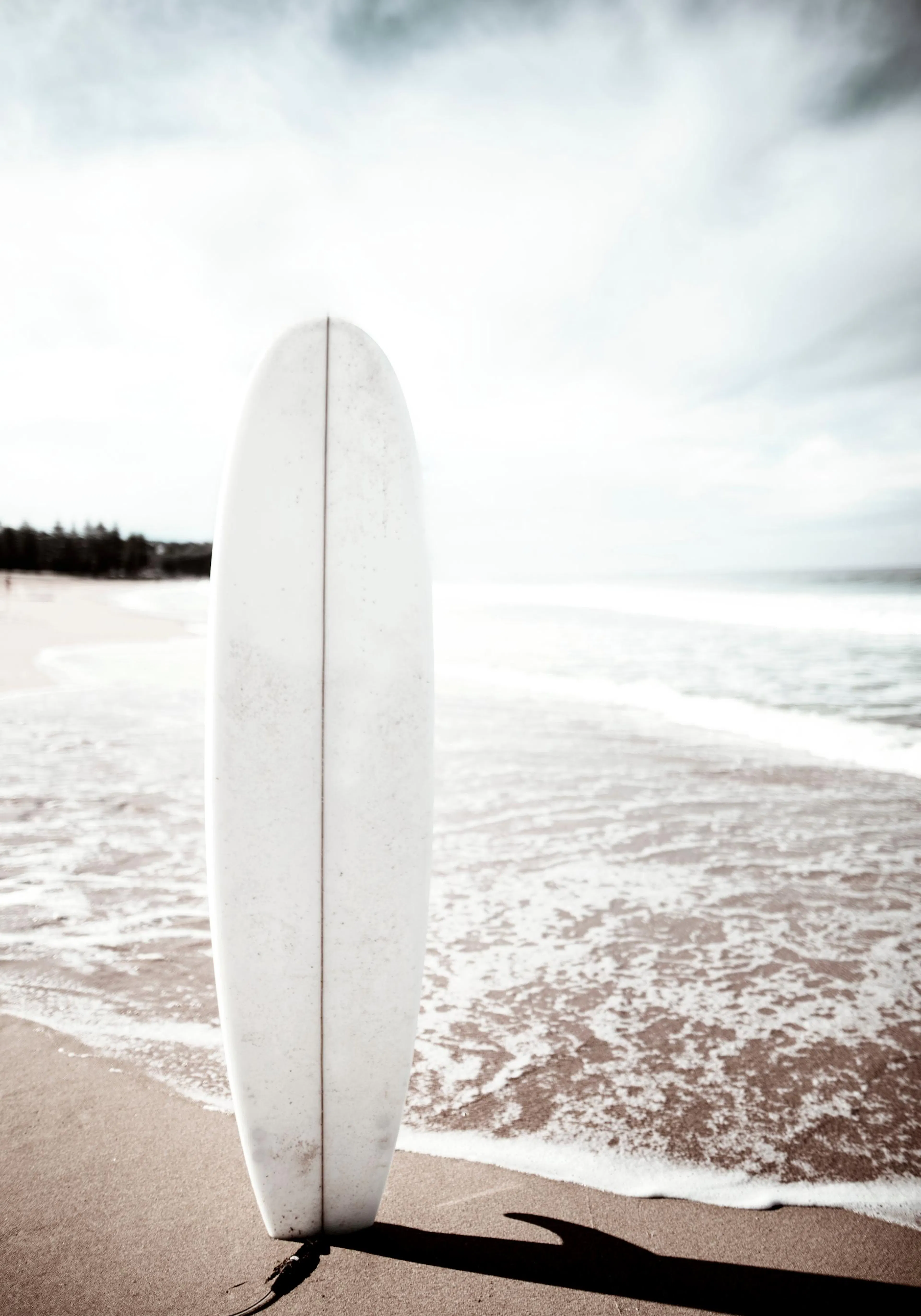 Een poster van een wit surfboard rechtop op het strand, met de oceaan en golven op de achtergrond.