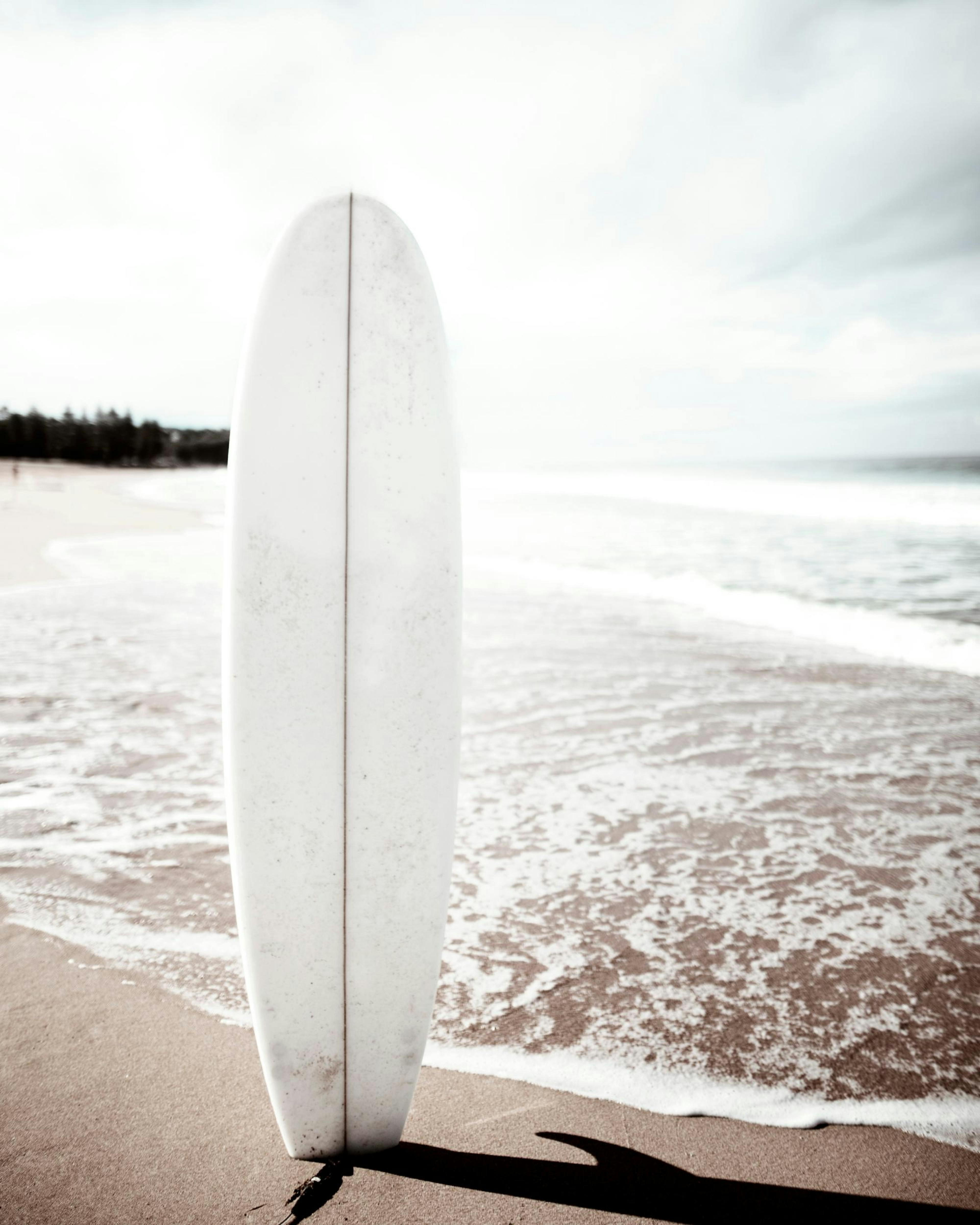 Póster de una tabla de surf blanca en la arena, con el océano y el cielo nublado de fondo.