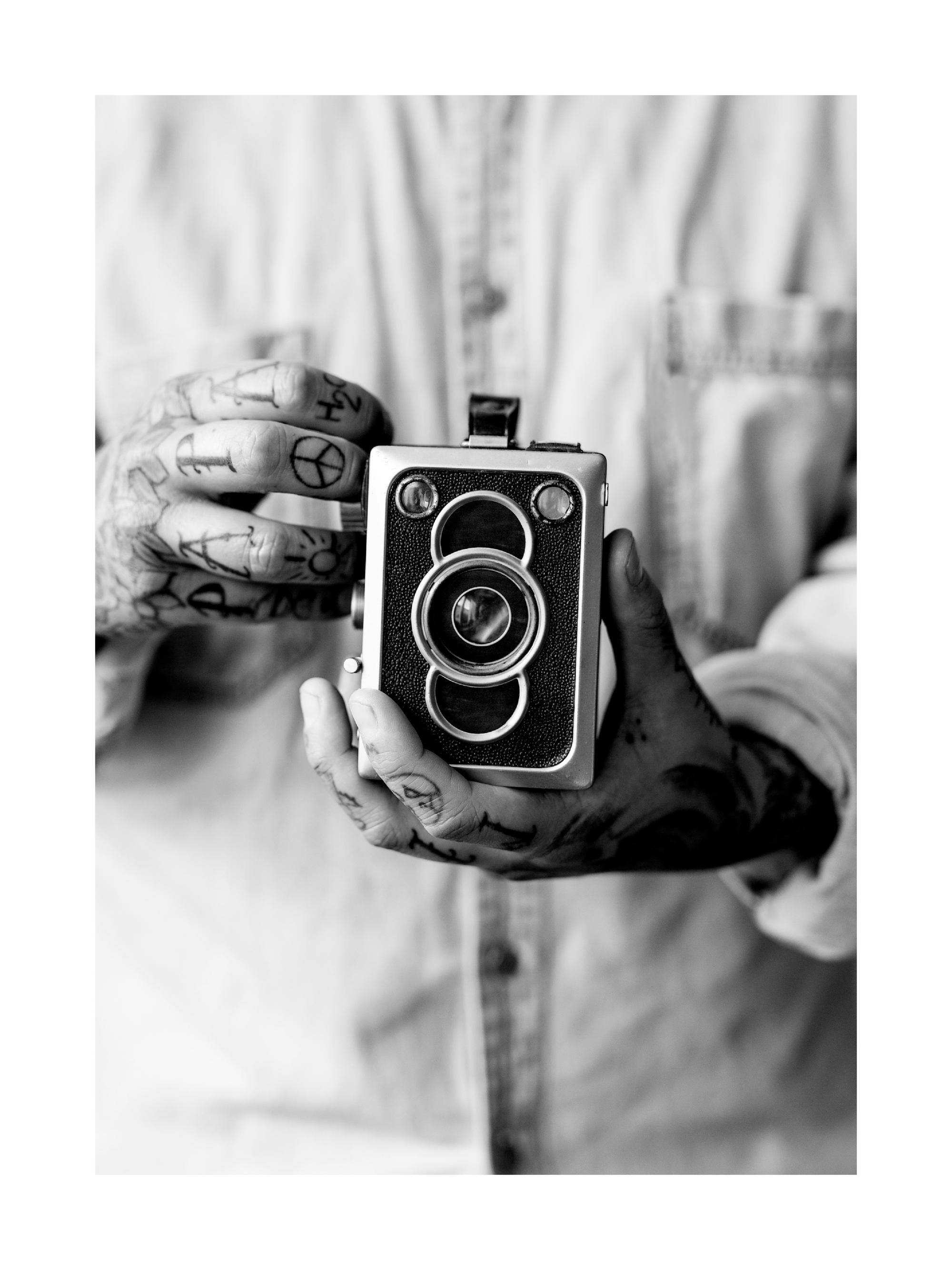 Mustavalkoinen juliste, jossa tatuoidut kädet pitelevät vanhaa kameraa. Kuva on lähikuva.