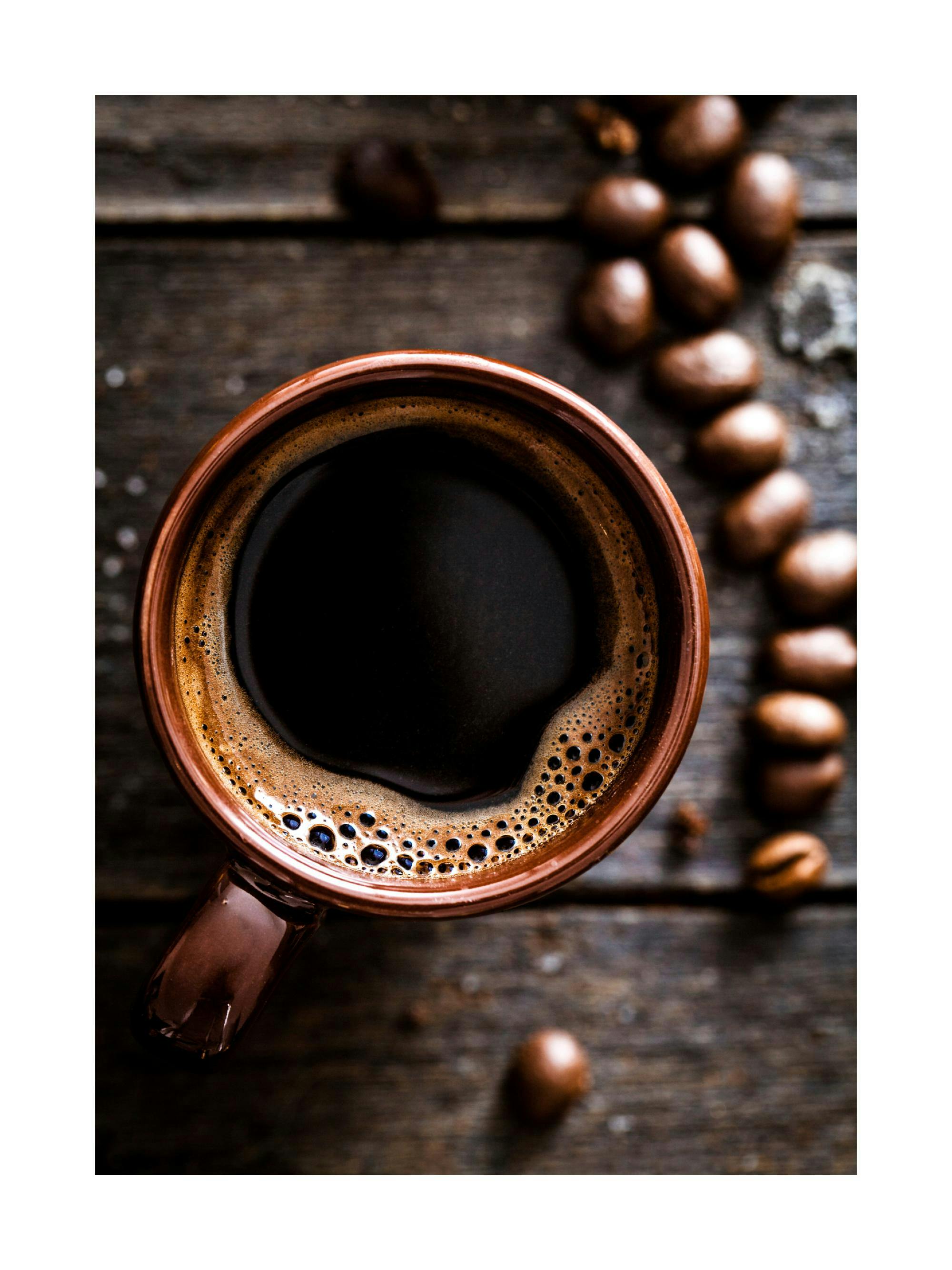 Plakat przedstawia kubek gorącej kawy z pianką i ziarna kawy na drewnianym tle.