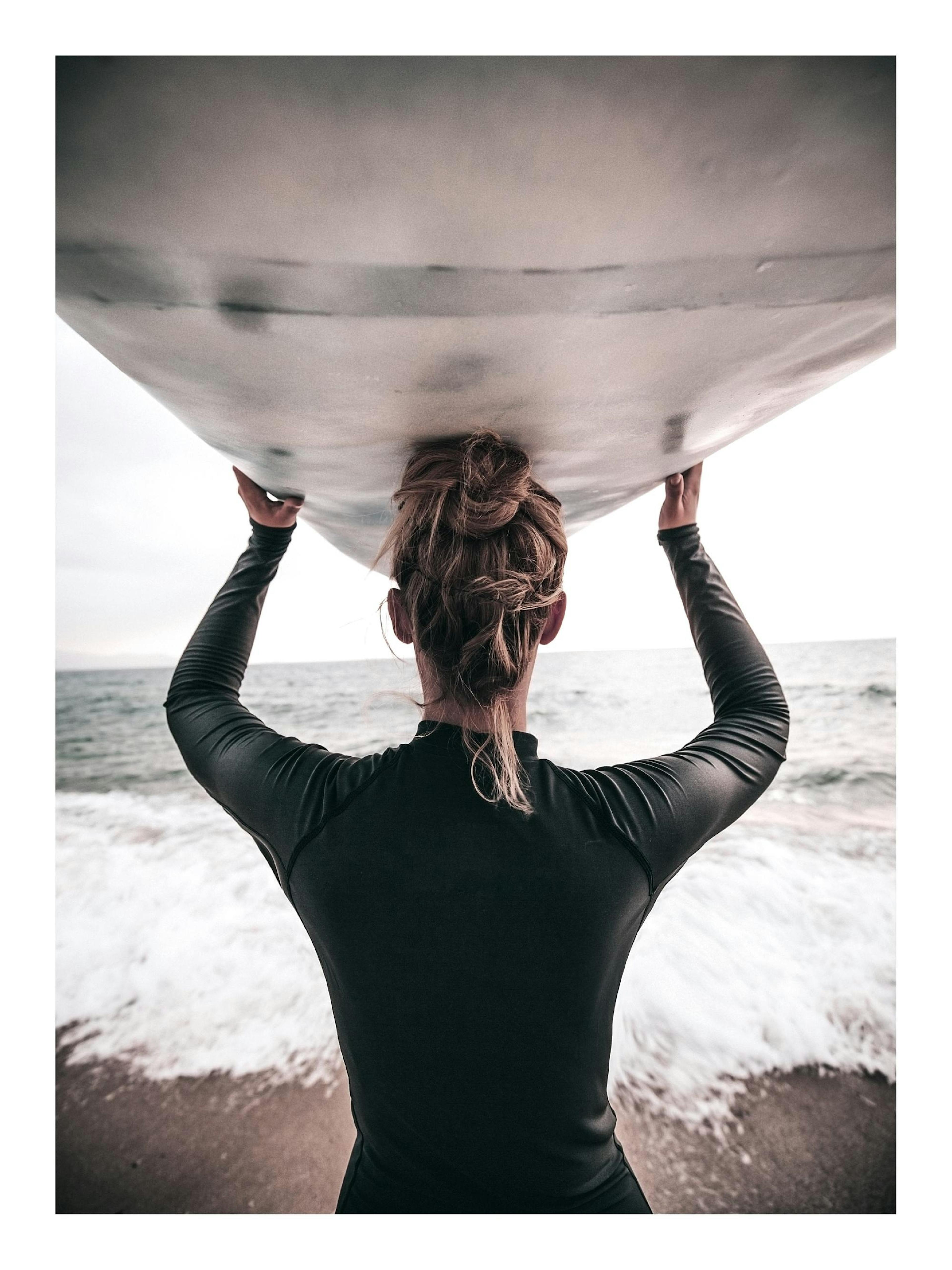 Un poster dune surfeuse en combinaison noire, portant sa planche sur la tête, face à locéan et aux vagues.
