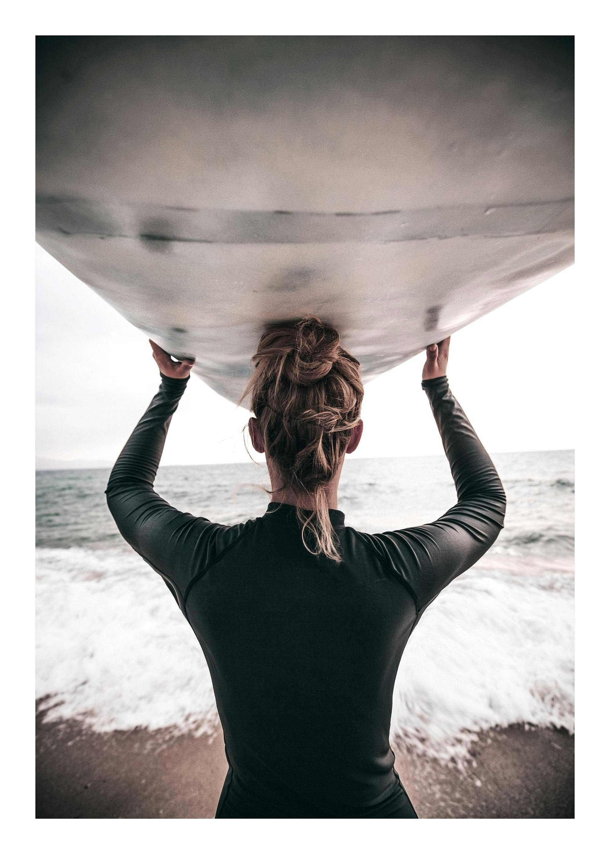 Een poster van een vrouw met donkere wetsuit die een surfboard op haar hoofd draagt op het strand, met de zee op de achtergrond.