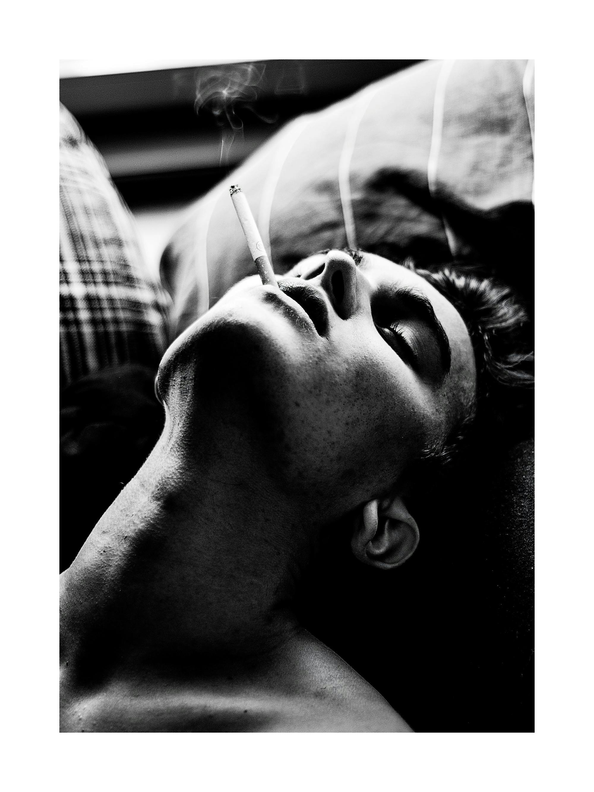 タバコをくわえて仰向けに横たわる男性の顔を捉えた白黒のポートレートポスター。煙が立ち上る。