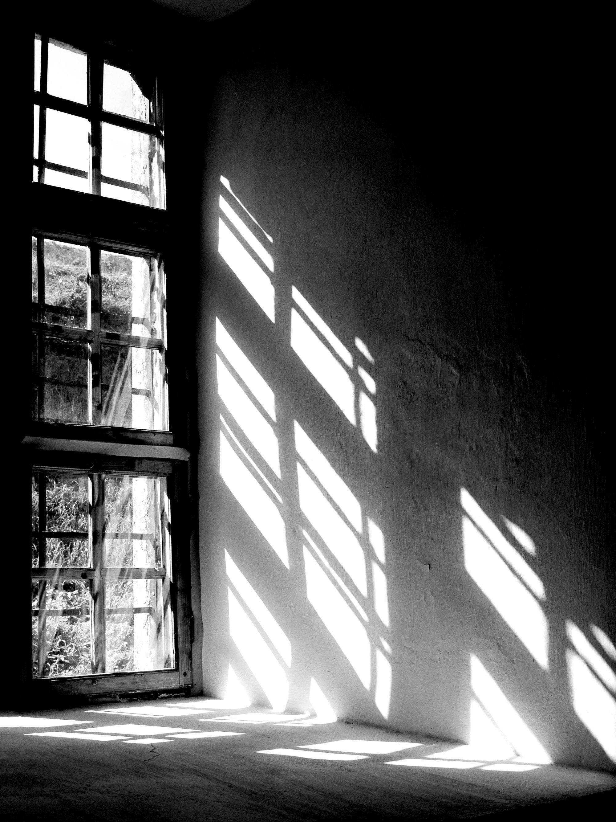 Schwarz-weiß Poster eines alten Fensters, das Sonnenlichtstreifen auf eine grobe Wand wirft und Schattenmuster erzeugt.