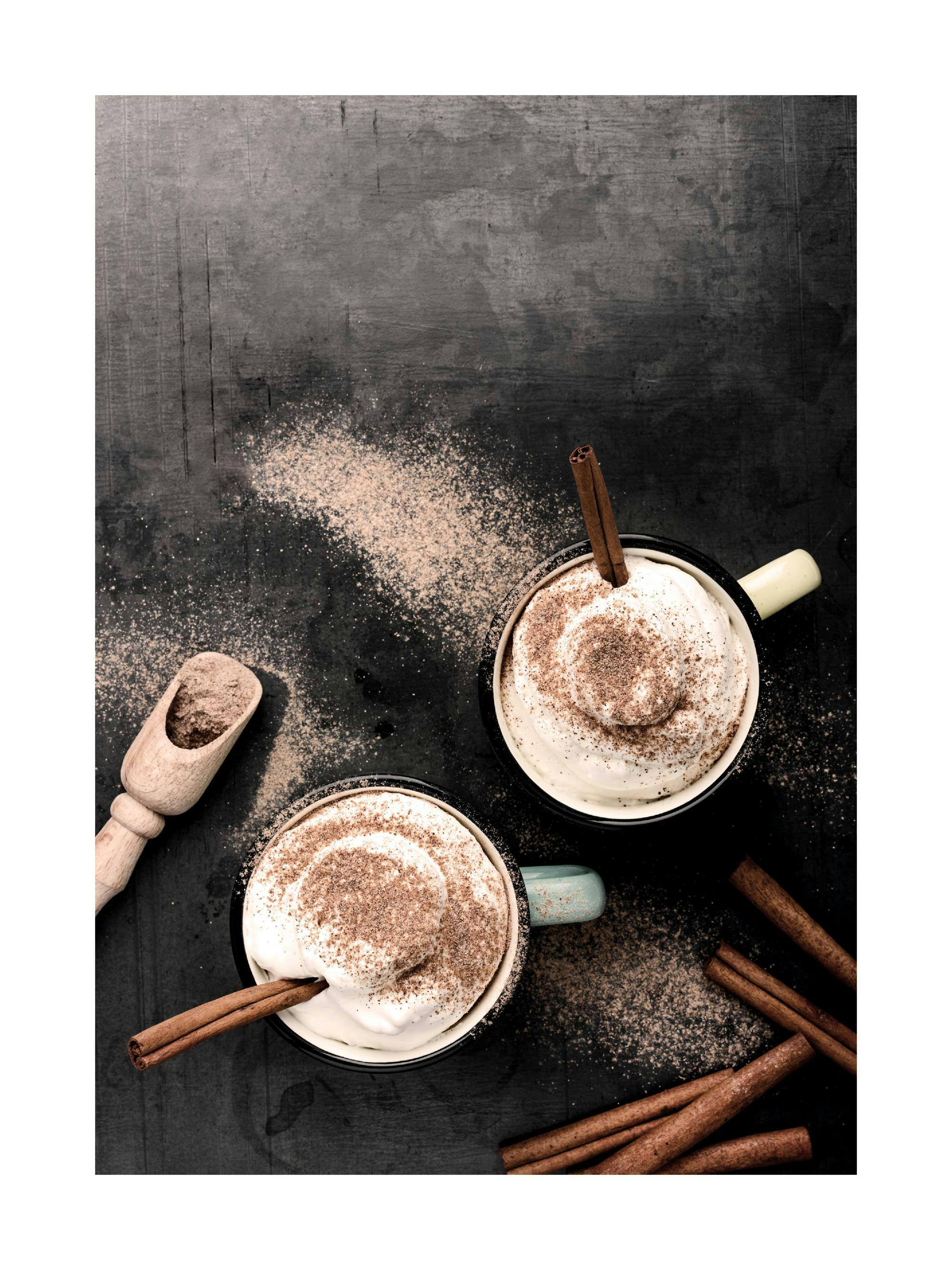 Un poster cu două căni de ciocolată caldă cu frișcă, presărate cu cacao și bețișoare de scorțișoară, pe o masă închisă la culoar