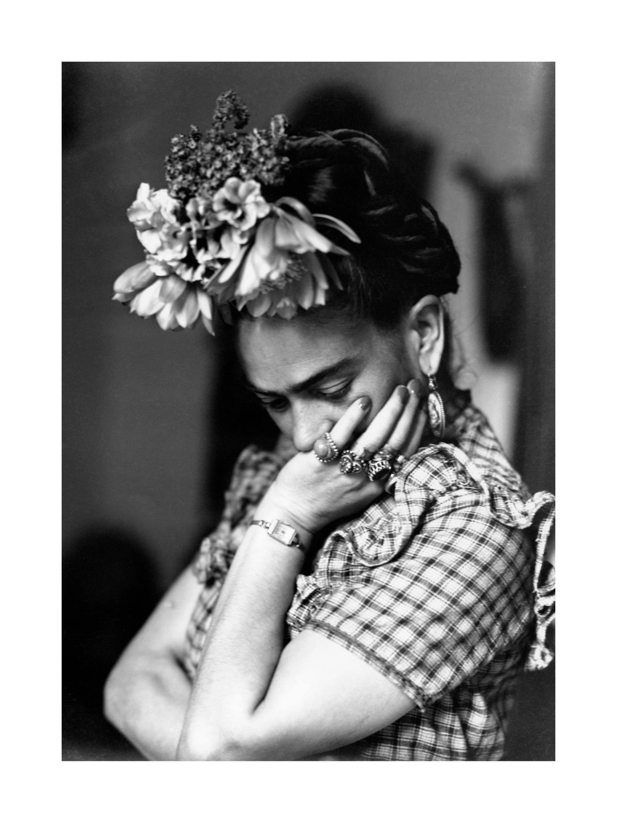 Poster alb-negru cu Frida Kahlo cu flori în păr, cotul sprijinit pe brațul încrucișat și mâna la bărbie.