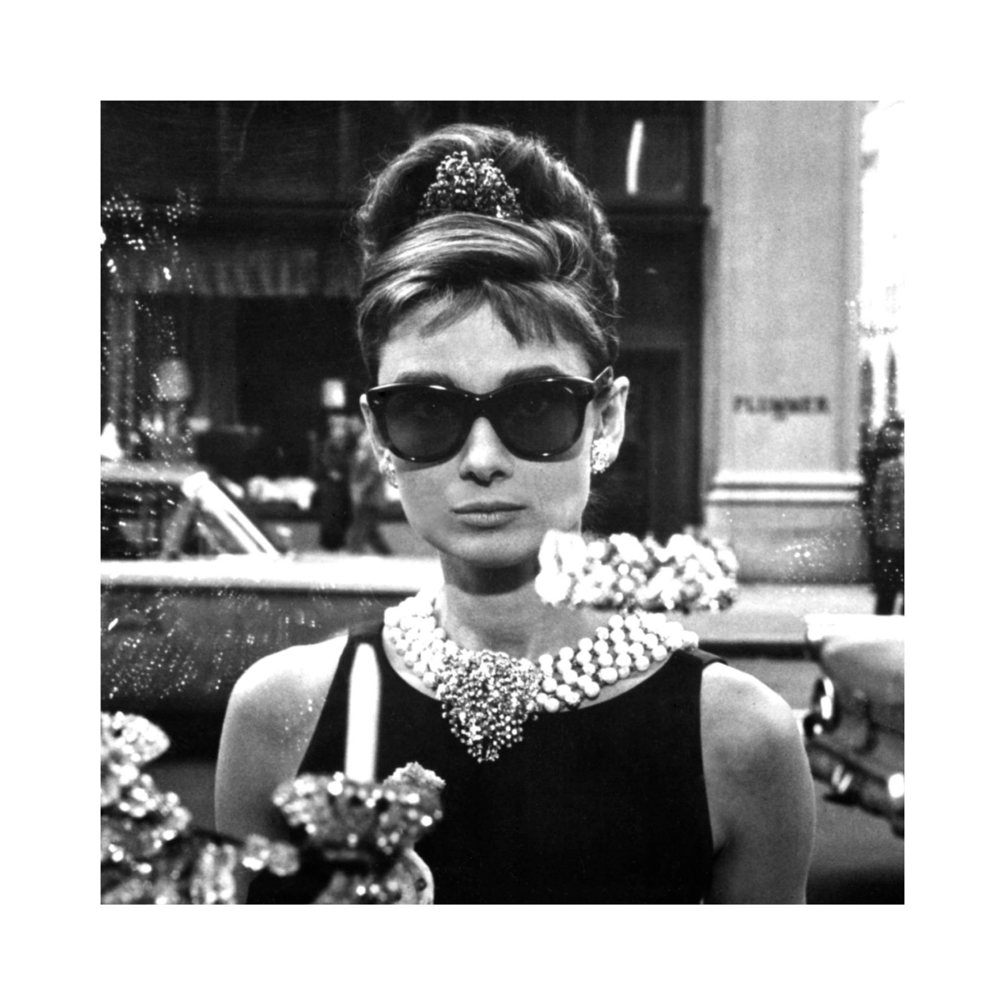 Zwart-wit poster van Audrey Hepburn als Holly Golightly met zonnebril en parelketting, kijkend door een etalage.