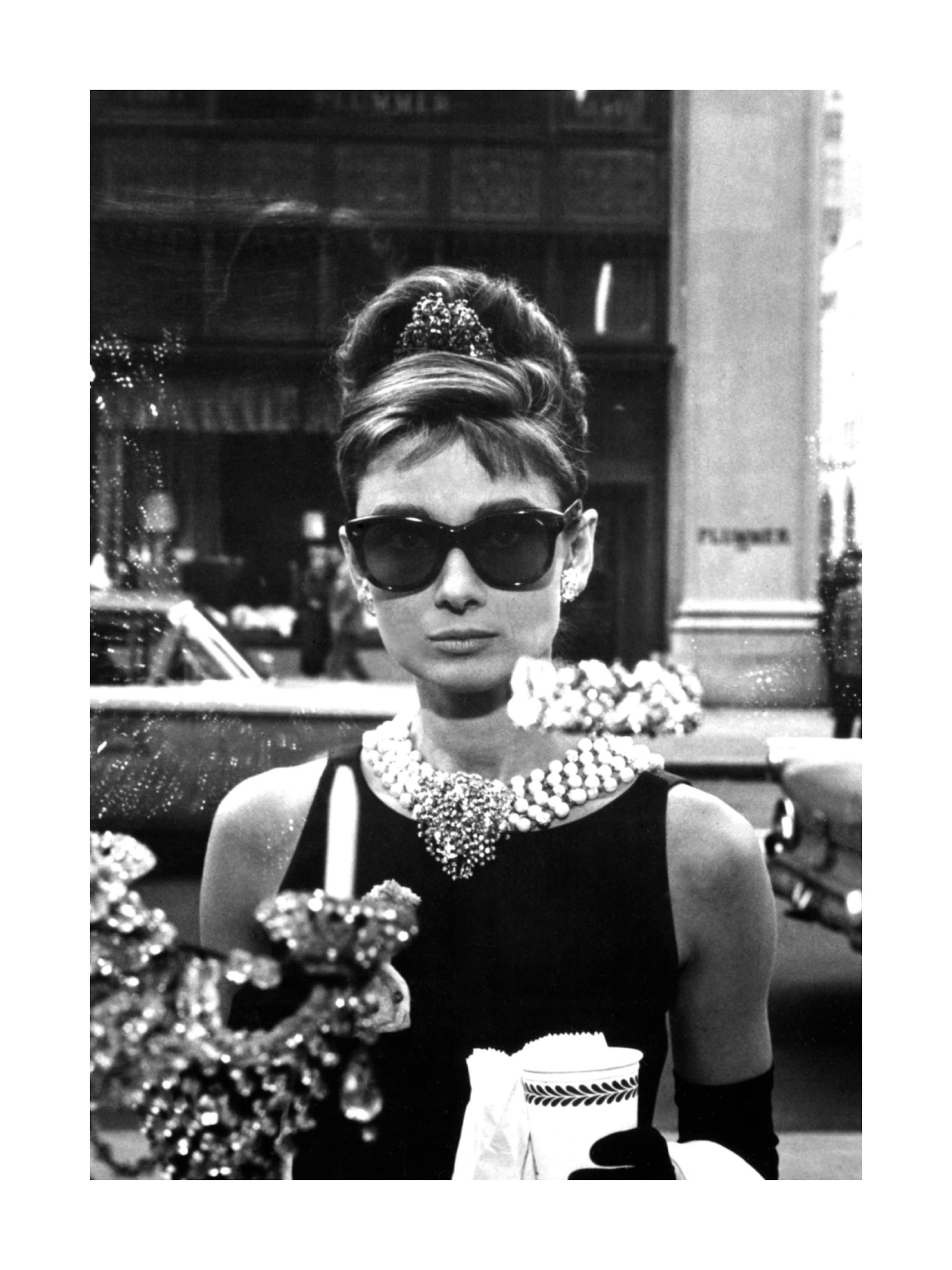 Póster en blanco y negro de Audrey Hepburn con gafas de sol, collar de perlas y tiara, mirando por un escaparate.