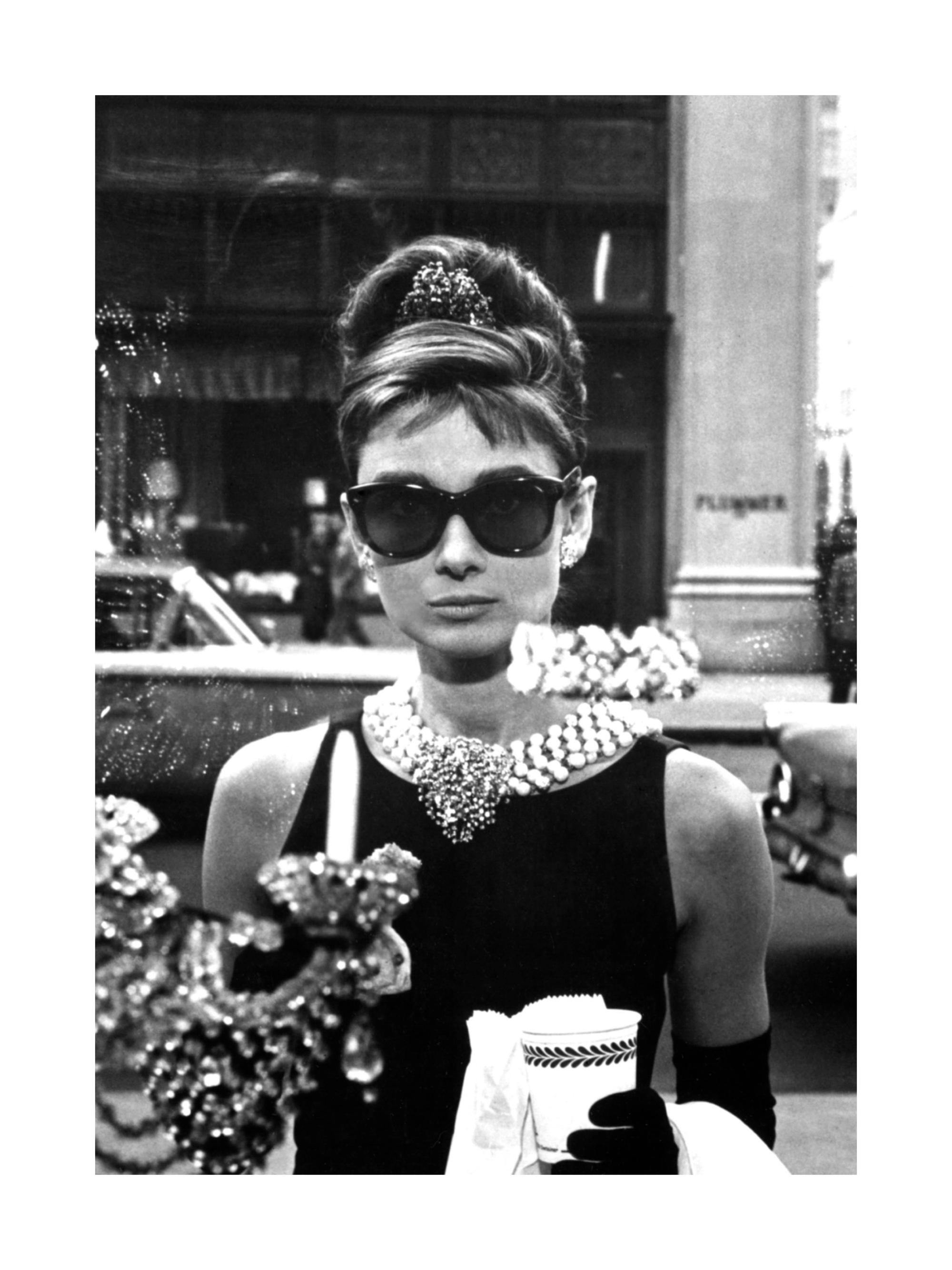 A black and white poster of a woman, Audrey Hepburn, wearing sunglasses and a black dress, holding a cup.