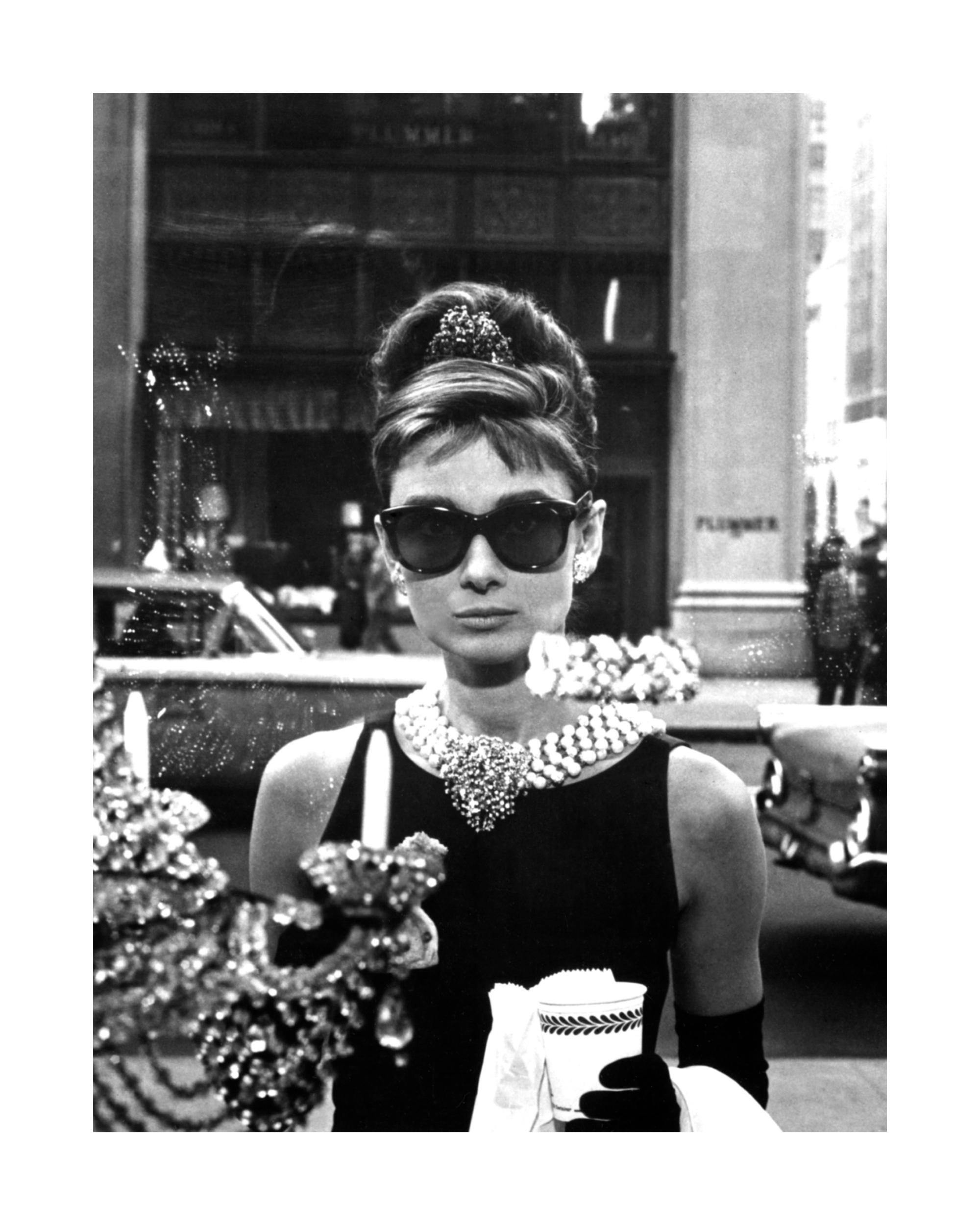 A black and white poster of Audrey Hepburn as Holly Golightly wearing sunglasses and a pearl necklace.