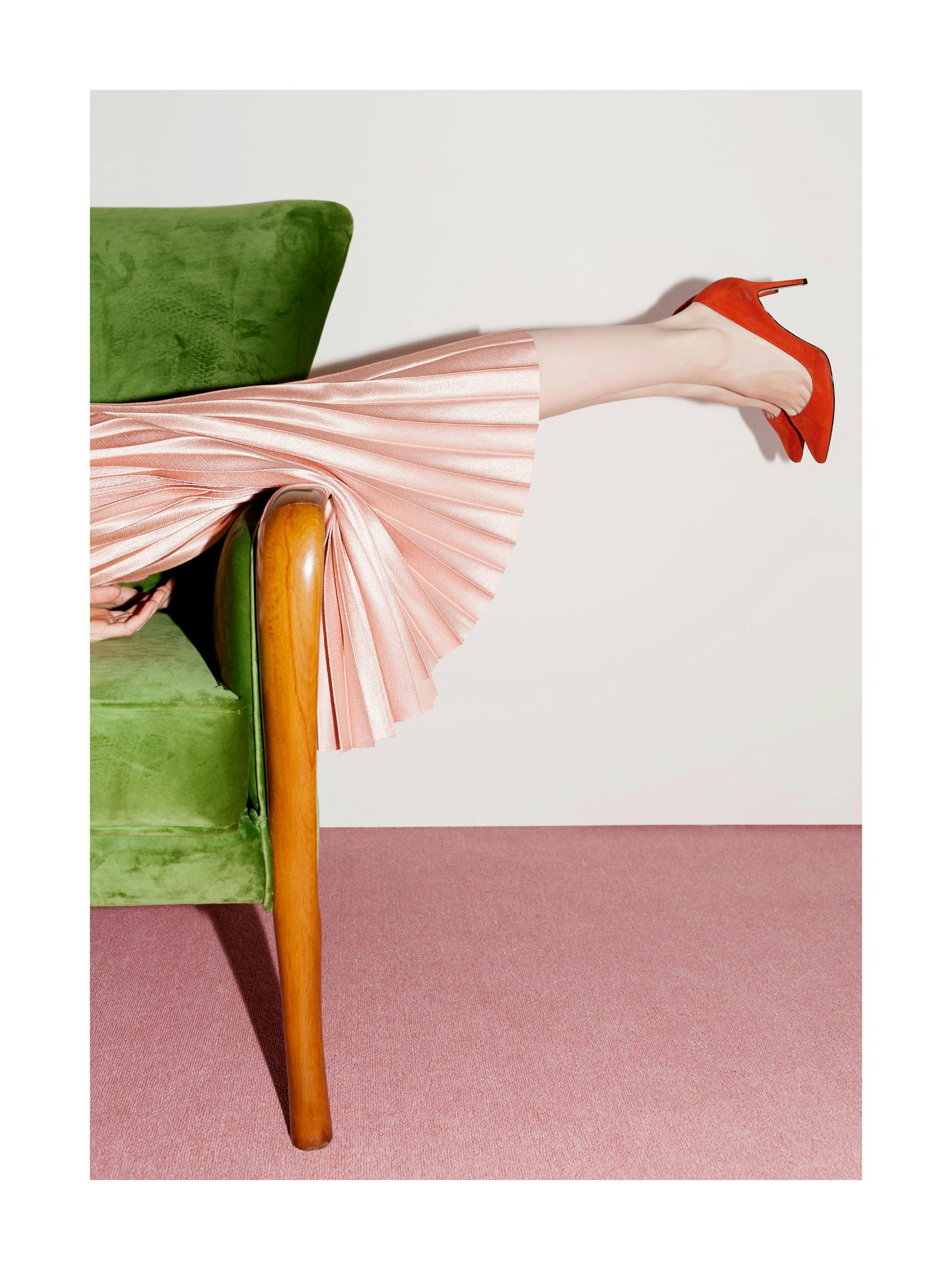 A poster featuring a persons legs in a pink pleated skirt and red high heels, resting on a green velvet chair.