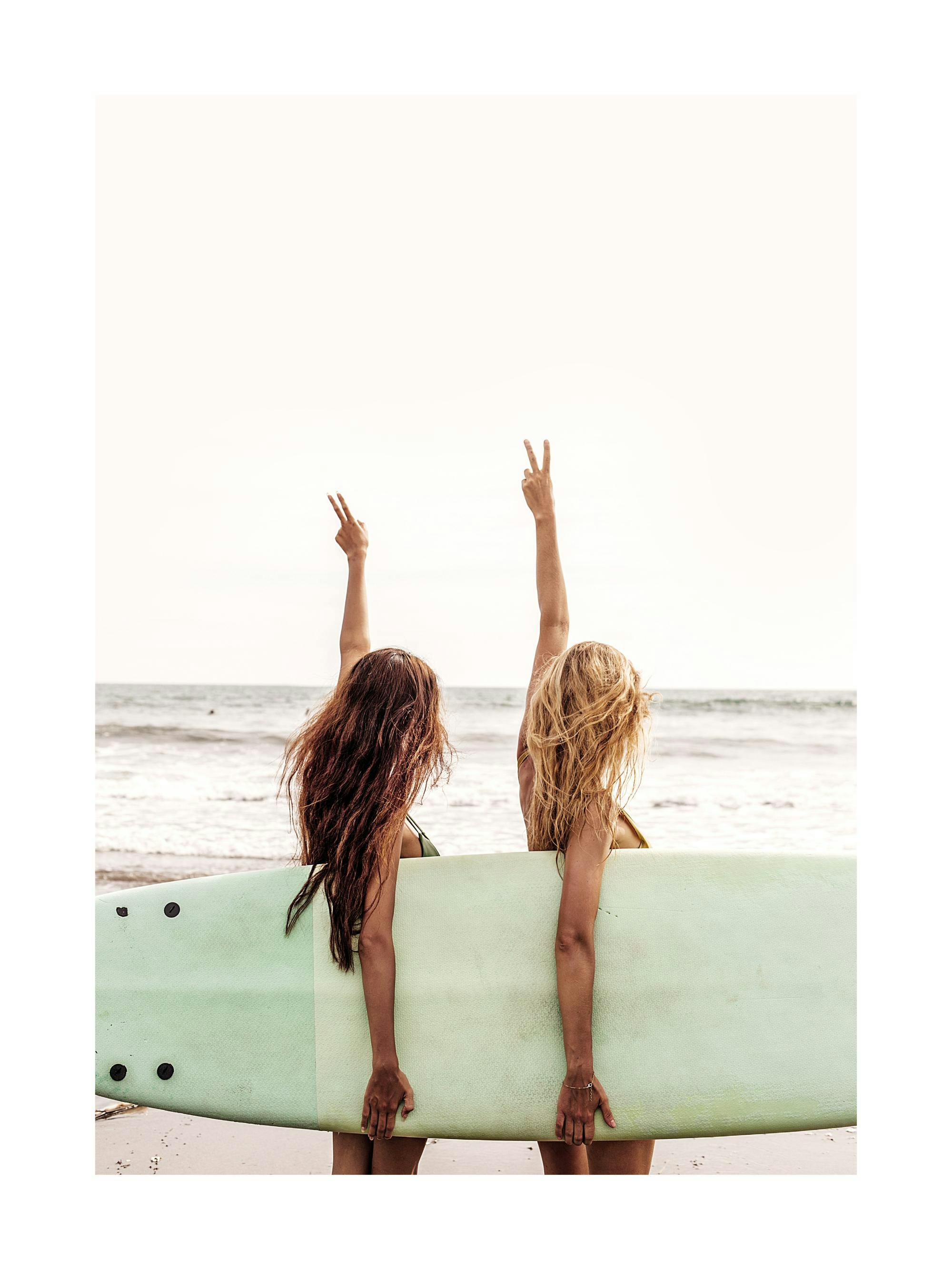 A poster of two women on a beach holding a surfboard, facing the ocean, with arms raised in peace signs.