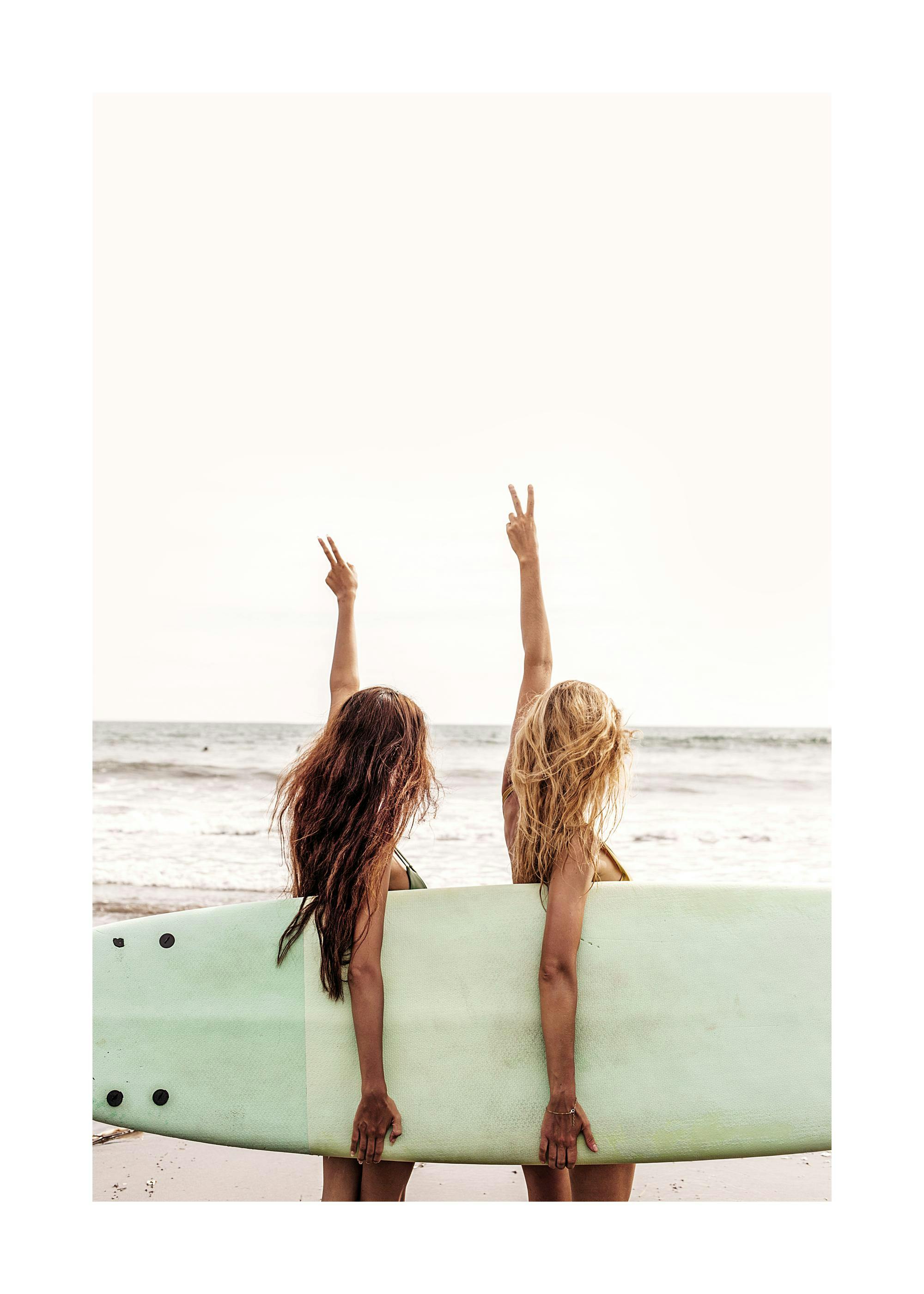 A poster featuring two women from behind, holding a surfboard on a beach, with arms raised showing peace signs.