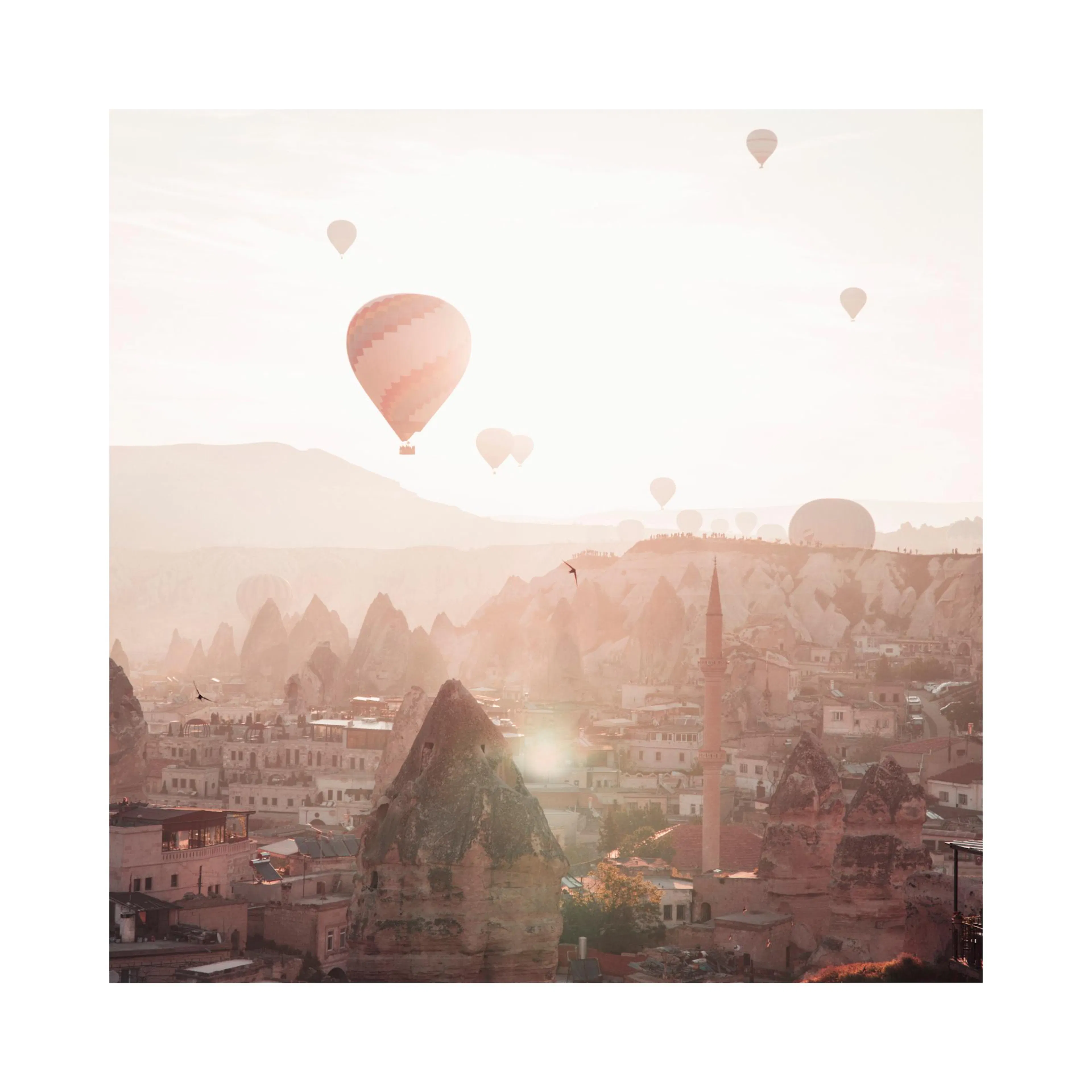 Un póster de un pintoresco paisaje de Capadocia con globos aerostáticos flotando sobre formaciones rocosas y casas al atardecer.