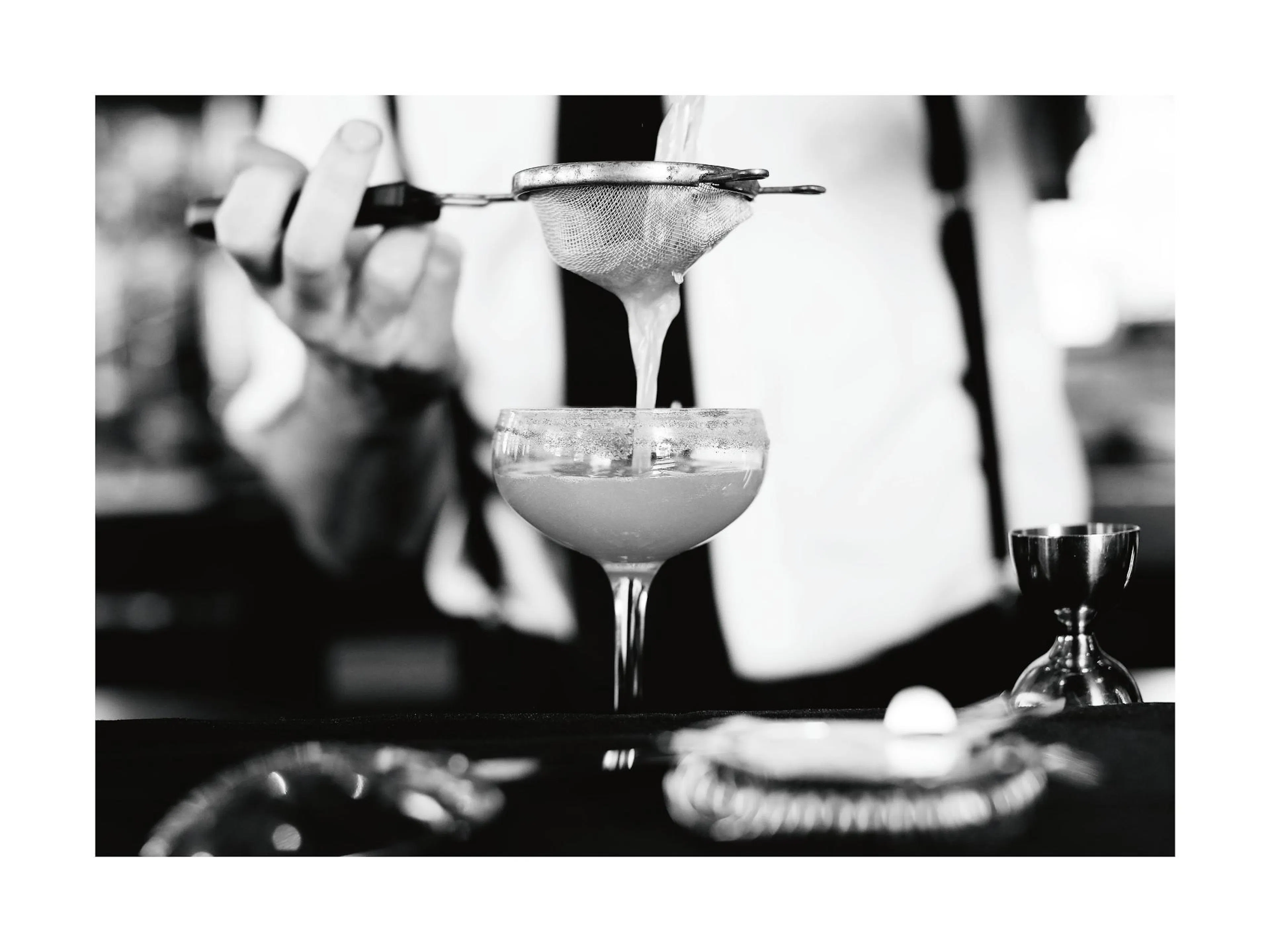 A black and white poster of a bartender straining a cocktail into a glass, with a jigger nearby.