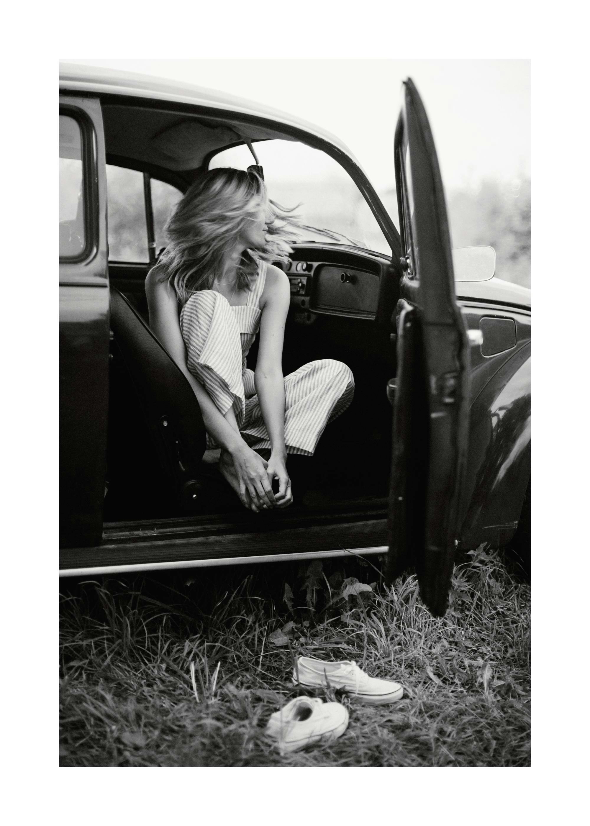 A black and white poster of a woman with wind-blown hair sitting in an old car with the door open, shoes on the ground.