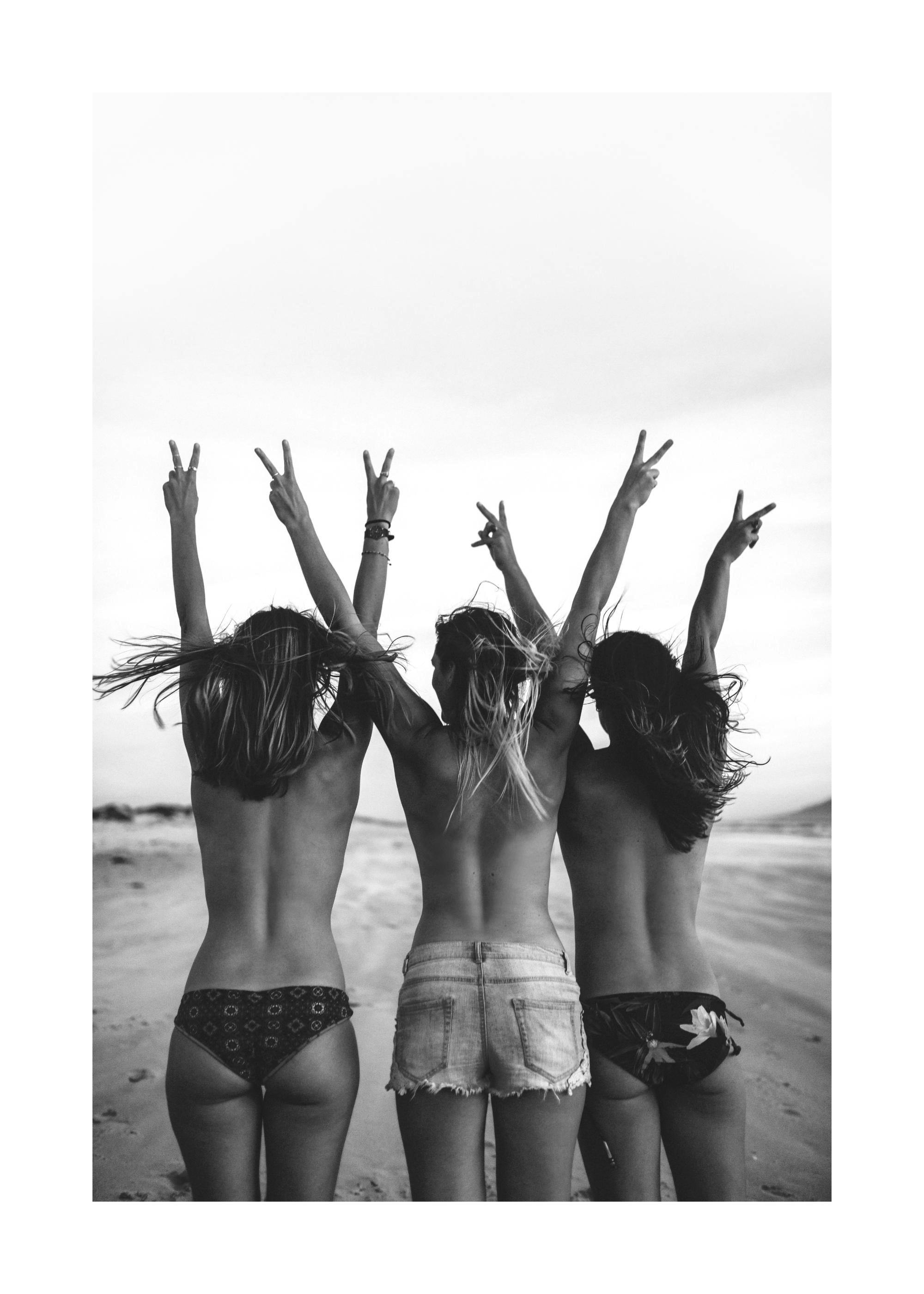 Póster en blanco y negro de tres mujeres de espaldas en la playa, con los brazos en alto haciendo la señal de la victoria.
