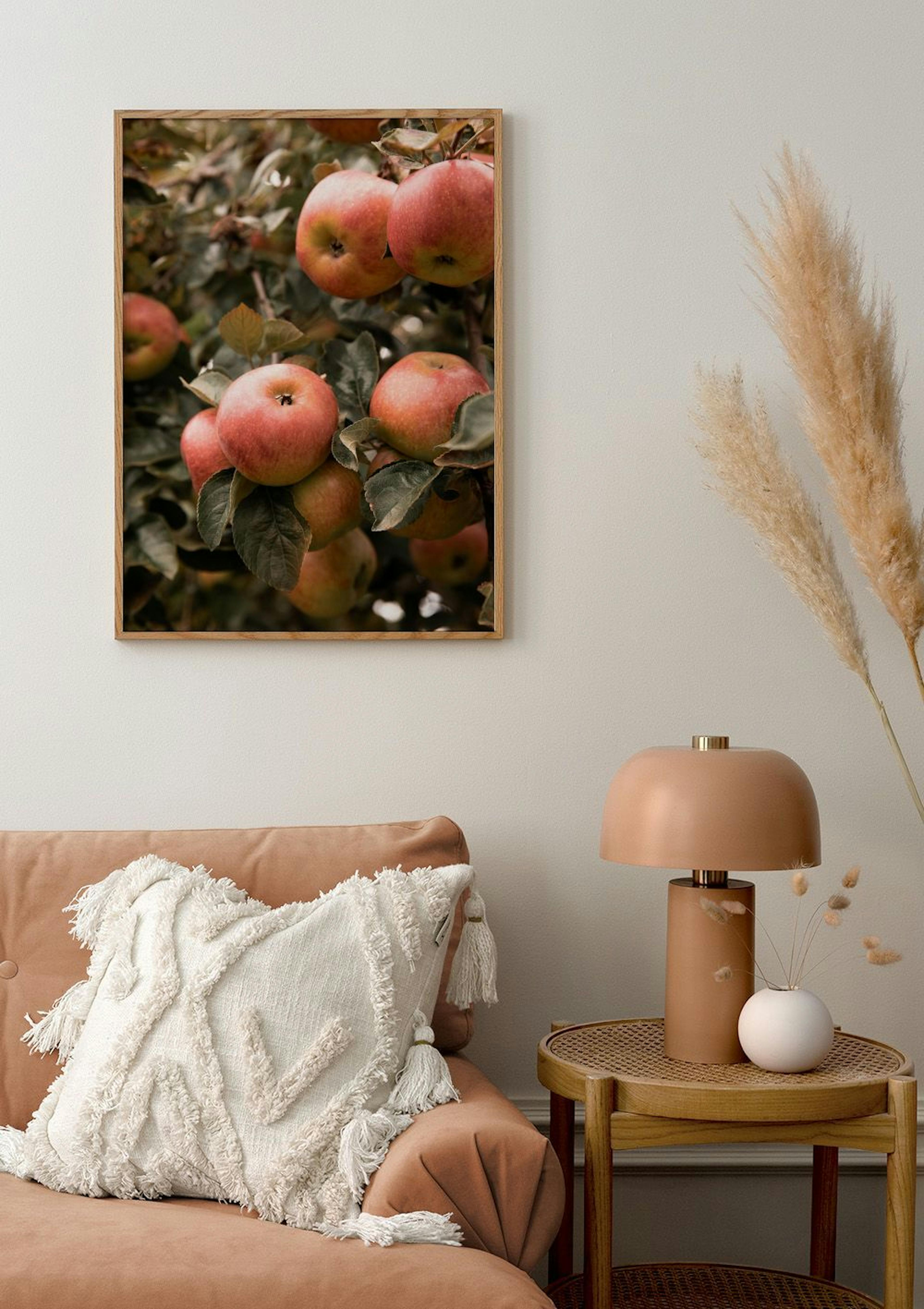 Poster of red apples on branches, hanging above a peach sofa with a white cushion, beside a lamp and dried pampas grass.