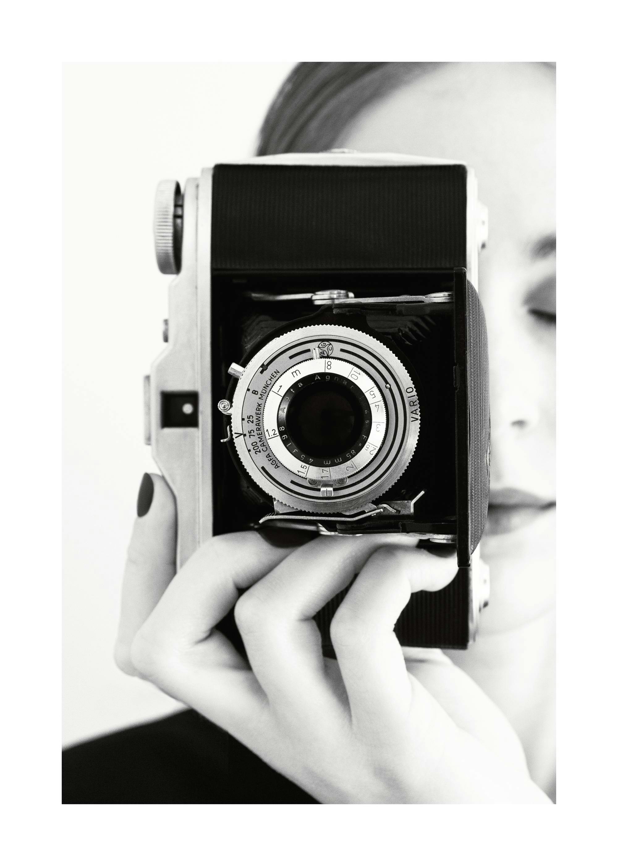 A black and white poster of a person holding a vintage camera, obscuring their face, with focus on the lens.
