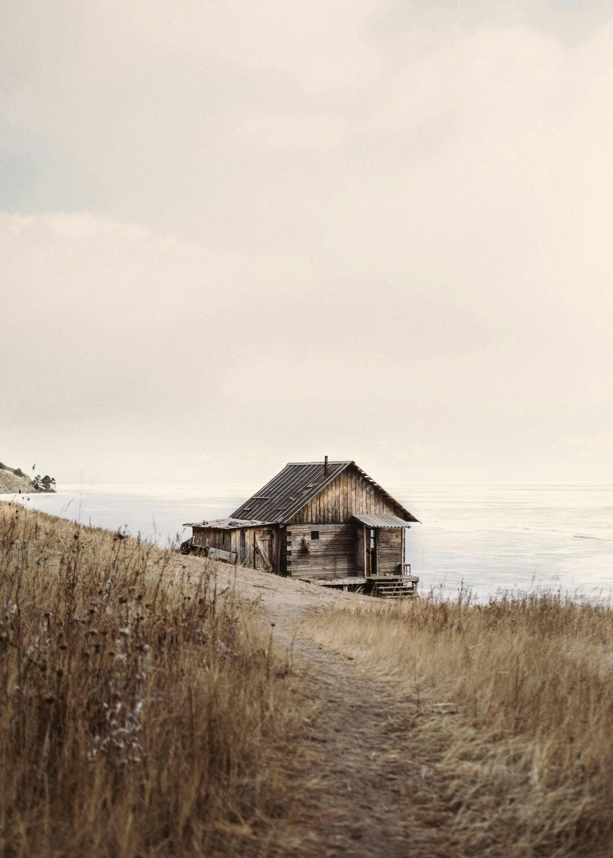 Poster: Eine einsame Holzhütte auf einem Hügel mit trockenem Gras, Blick auf ein Gewässer unter einem hellen Himmel.