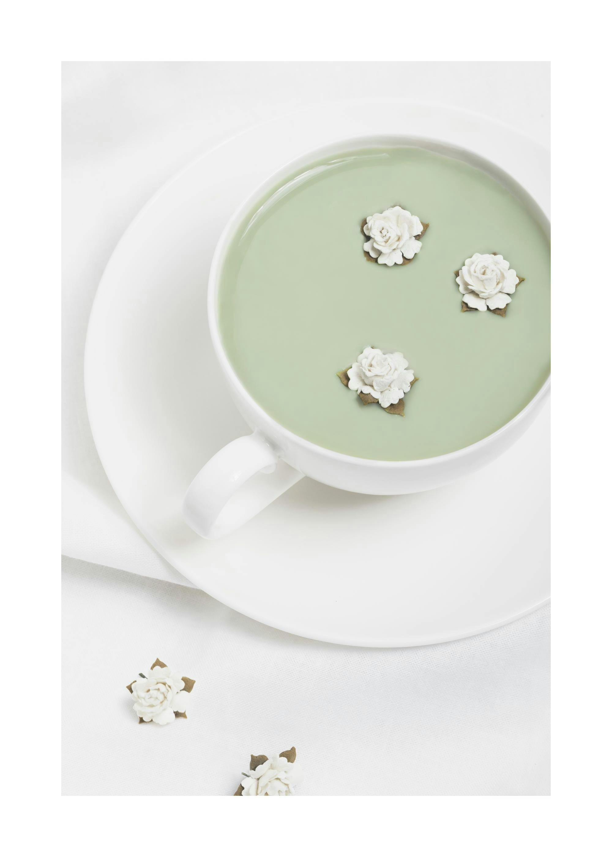 Póster de una taza de matcha con tres flores blancas flotando, sobre un plato blanco y un mantel blanco.