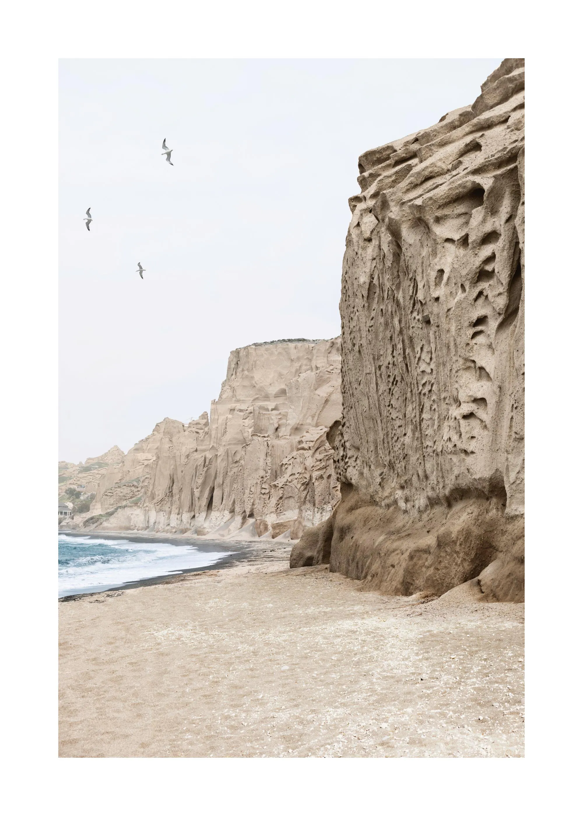 En poster som visar en strand med sand och mörka stenar, höga eroderade klippor och tre flygande måsar.