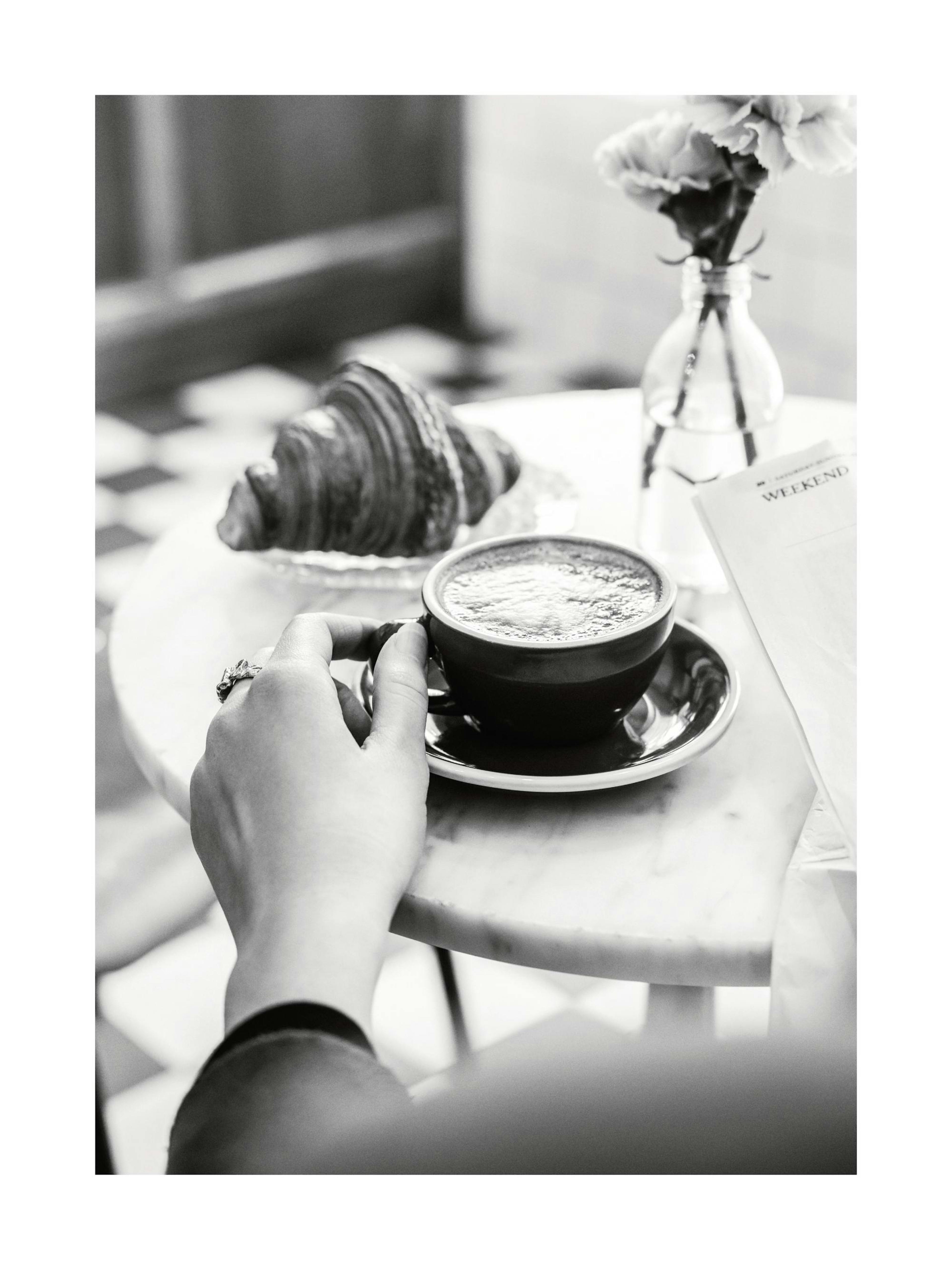 Póster en blanco y negro de una mano sosteniendo una taza de café, con un cruasán y flores en una mesa de mármol.