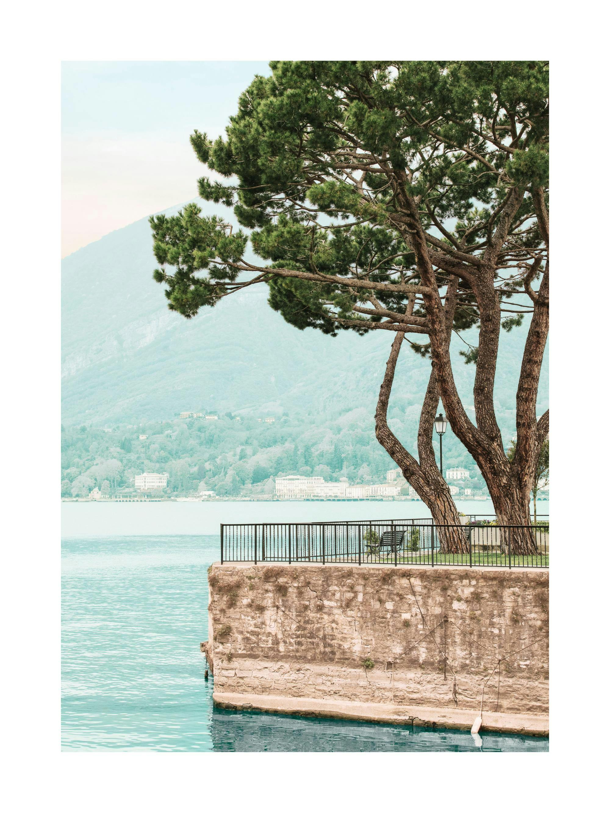 Poster darbres à feuilles persistantes sur un quai en pierre au bord dun lac turquoise, avec des montagnes en arrière-plan.