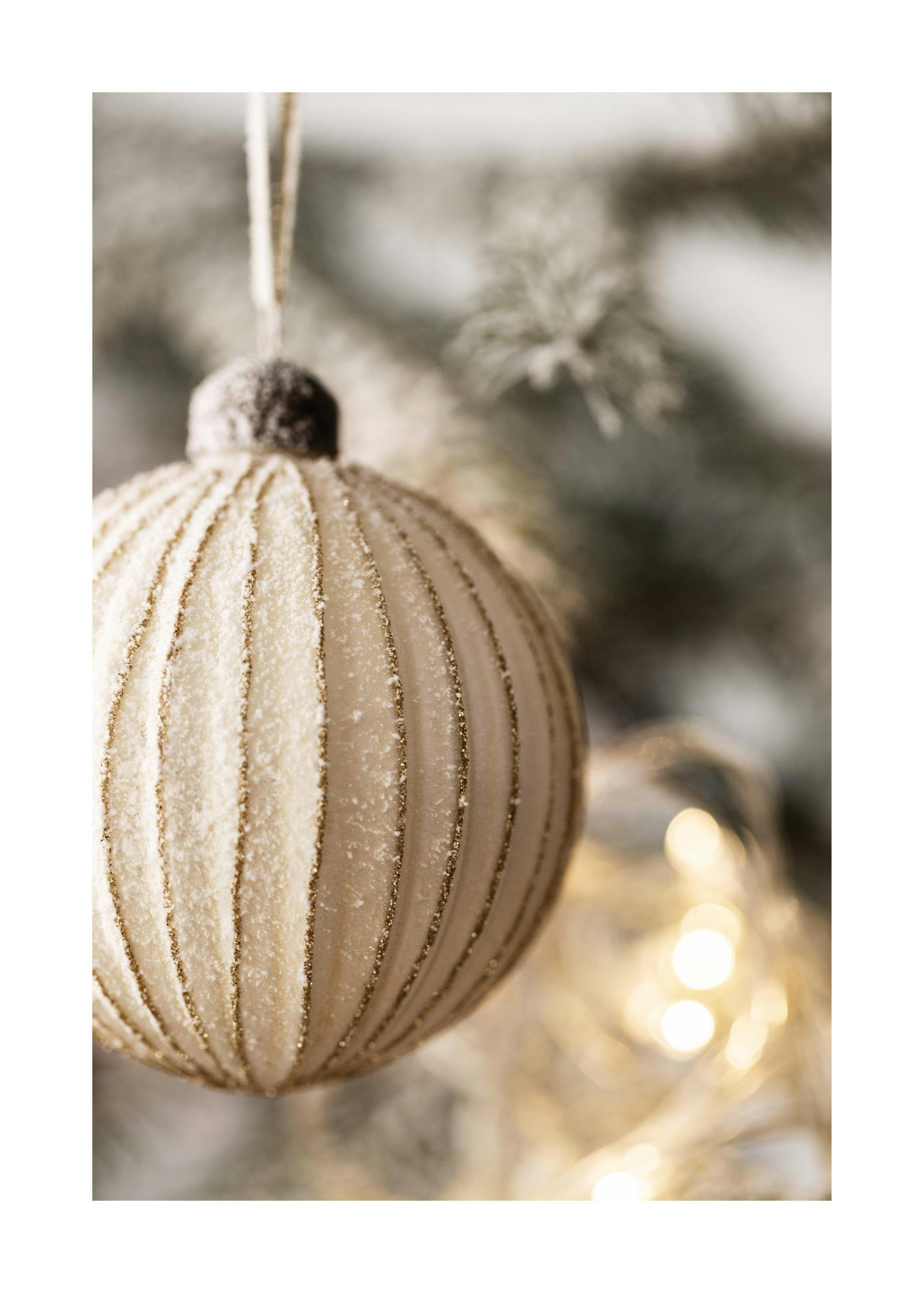 Un poster di una palla di Natale bianca e oro con glitter appesa a un albero di Natale innevato, con luci sfocate sullo sfondo.