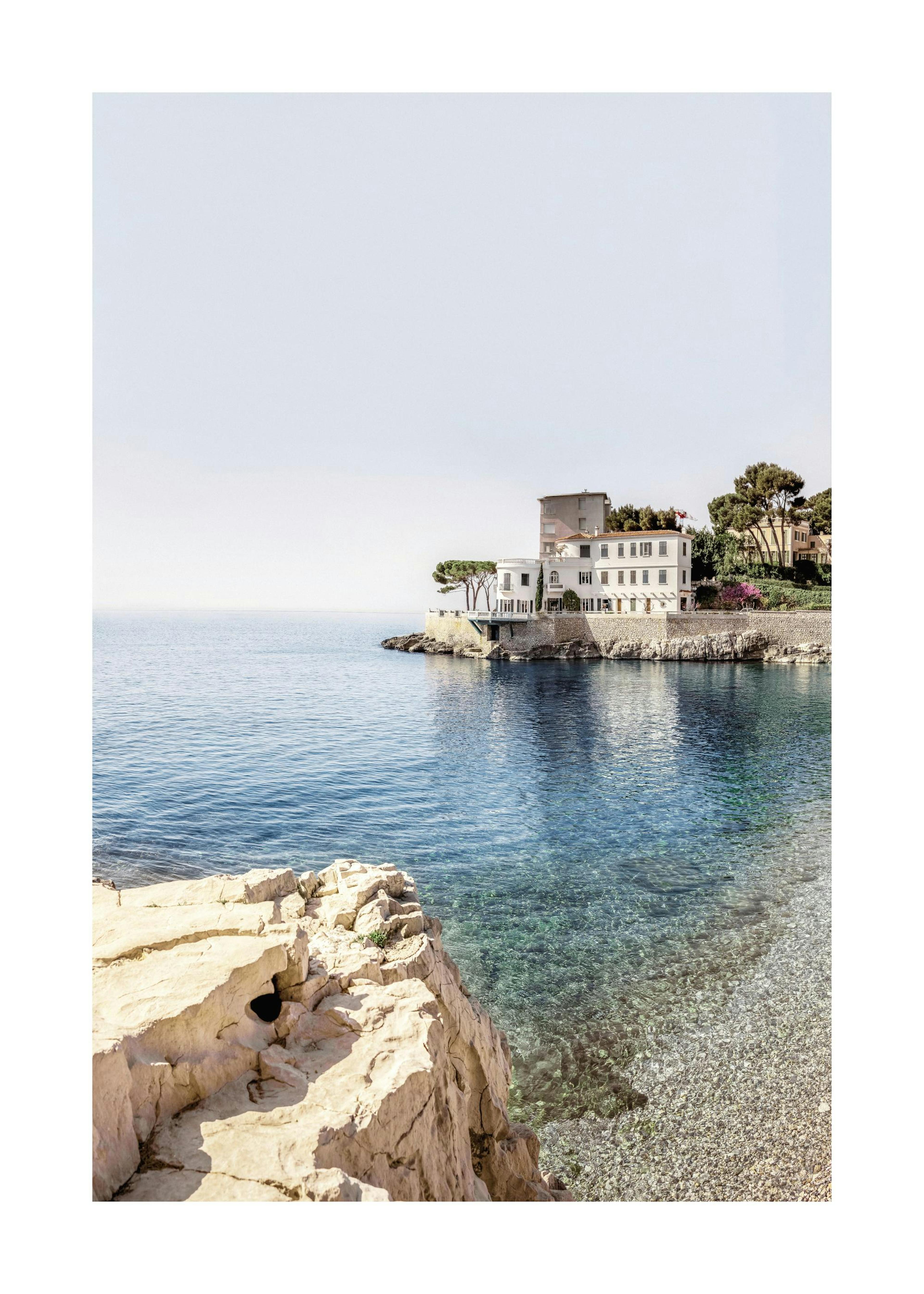 A poster featuring a Mediterranean coastal scene with a white building on a rocky promontory, clear blue water, and a pebbly bea