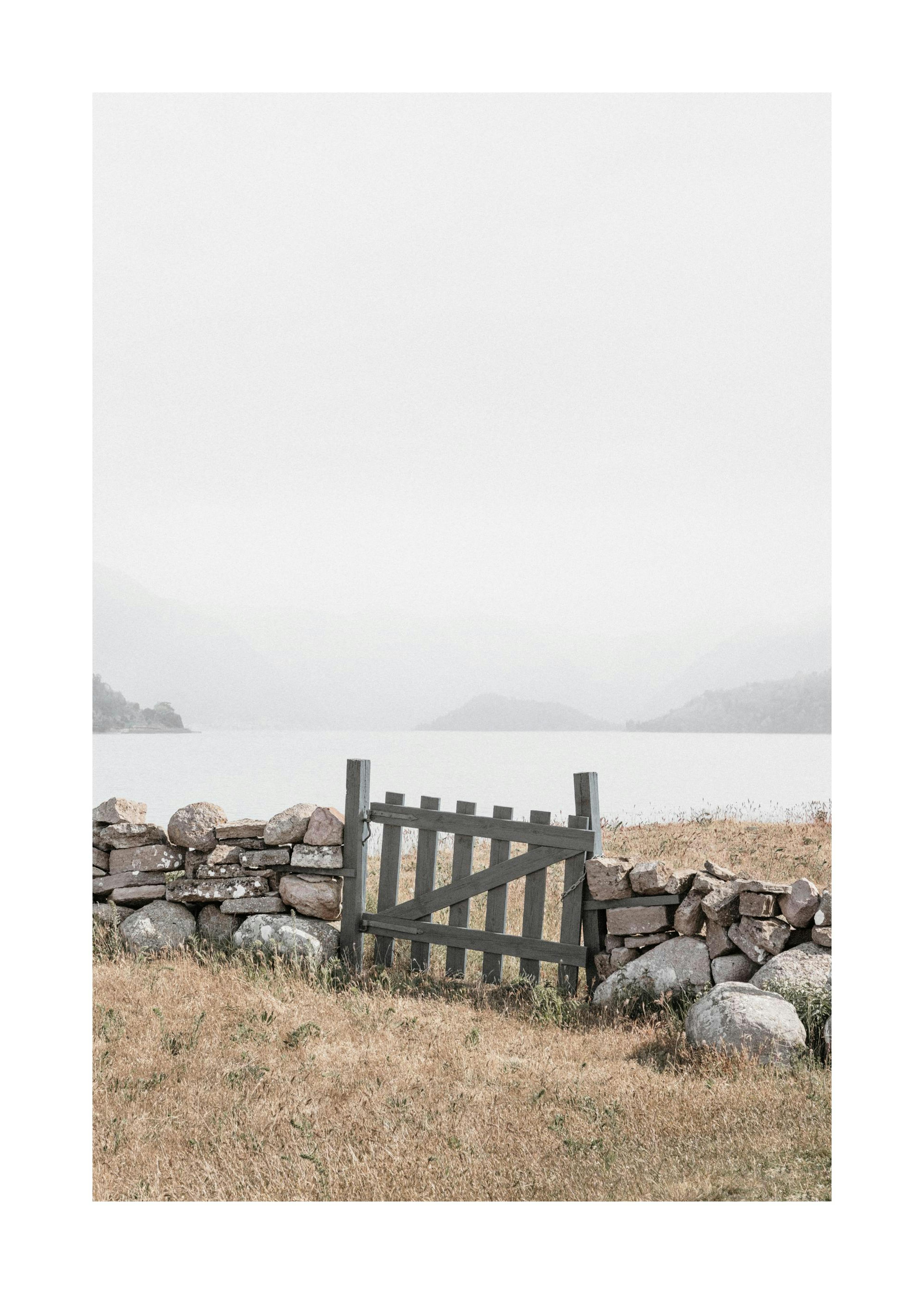 A poster featuring a weathered wooden gate within a stone wall, overlooking a lake with hazy mountains in the distance.