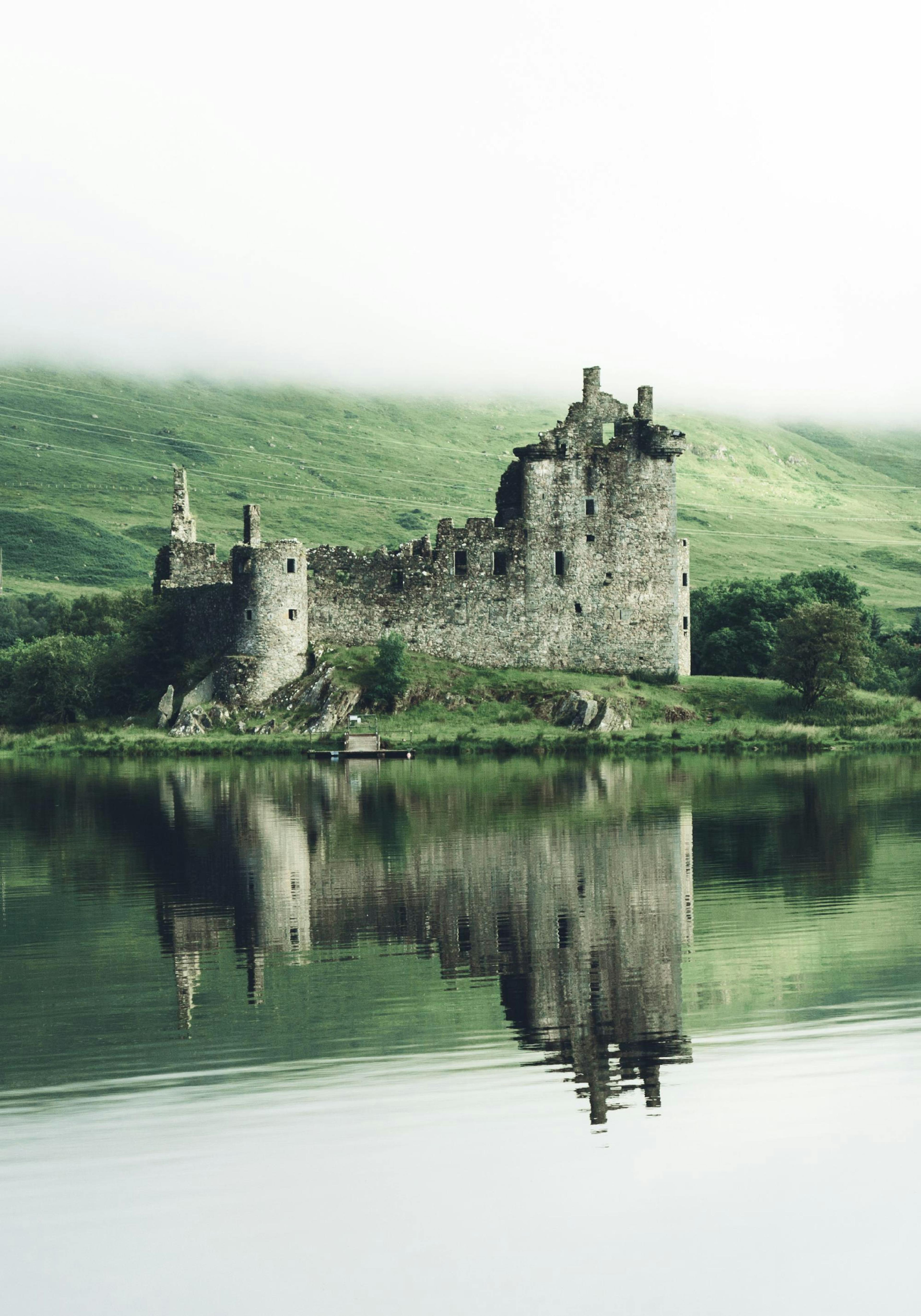 Poster: Historische Burgruine am Seeufer mit nebligen Bergen und Wasserspiegelung in Schottland oder Irland.