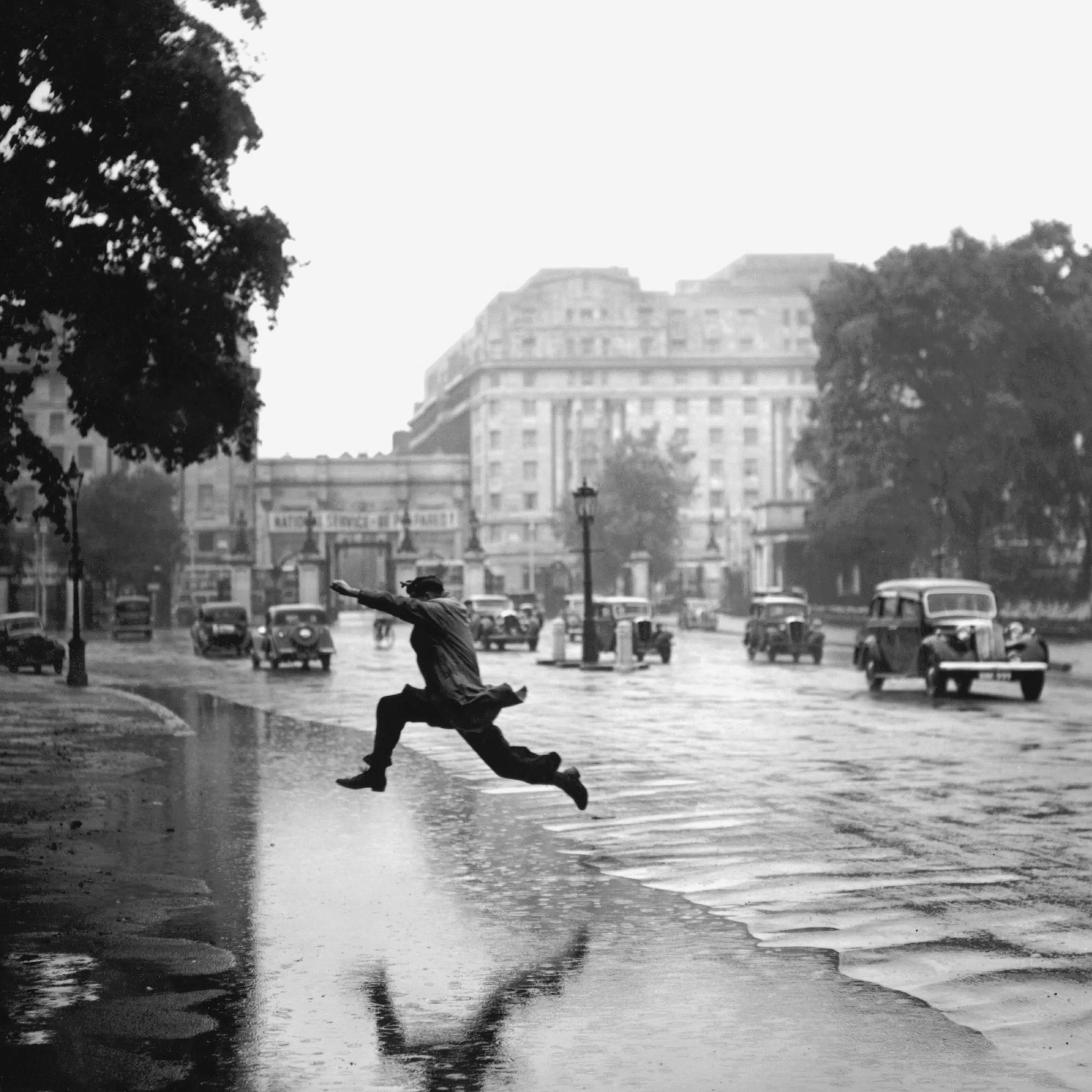 Poster: Schwarz-weißes Foto eines Mannes, der über eine Pfütze auf einer nassen Straße springt, mit Gebäuden und Autos im Hinter