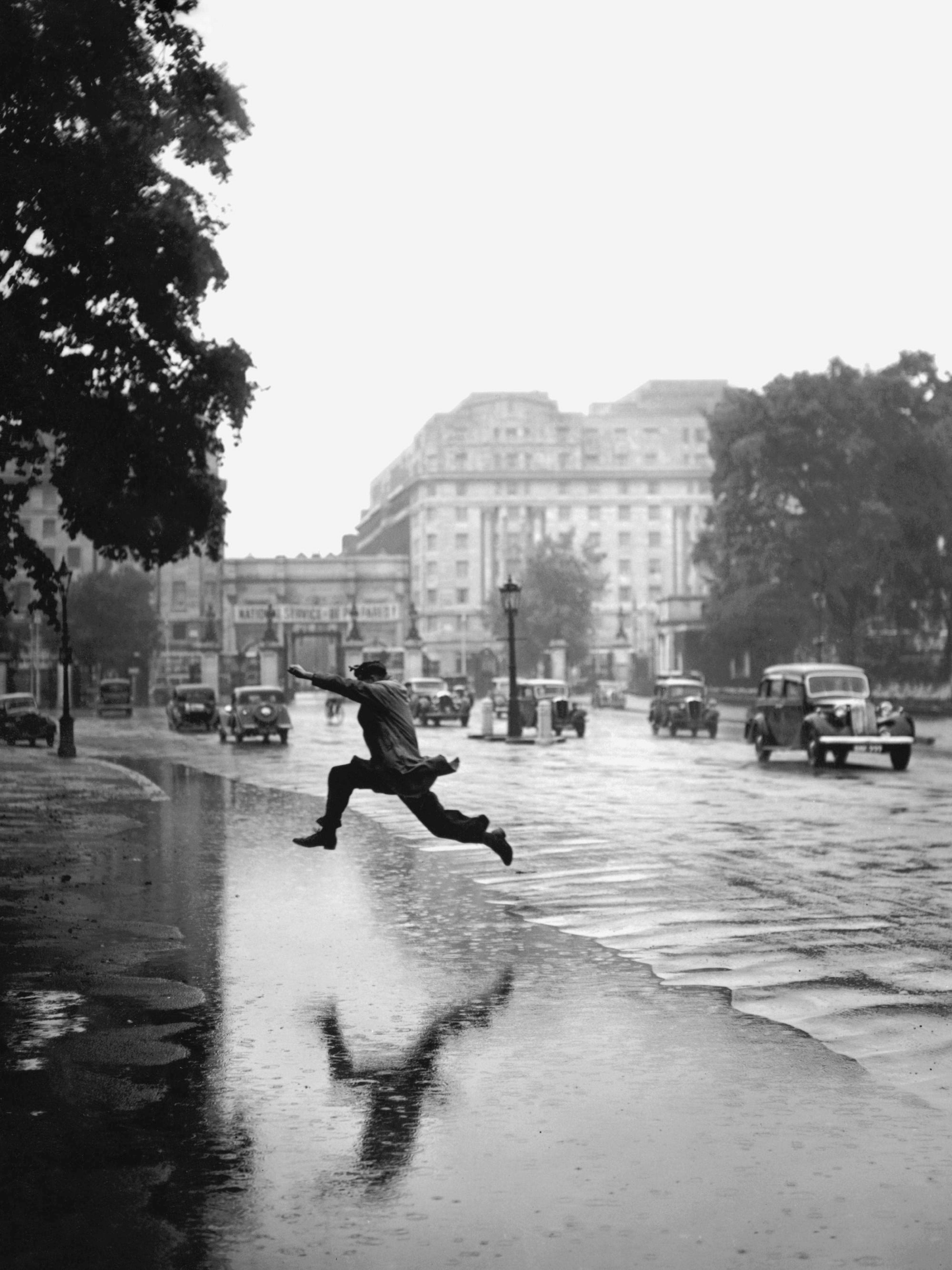 Póster en blanco y negro de un hombre saltando sobre un charco en una calle mojada, con coches y edificios a lo lejos.