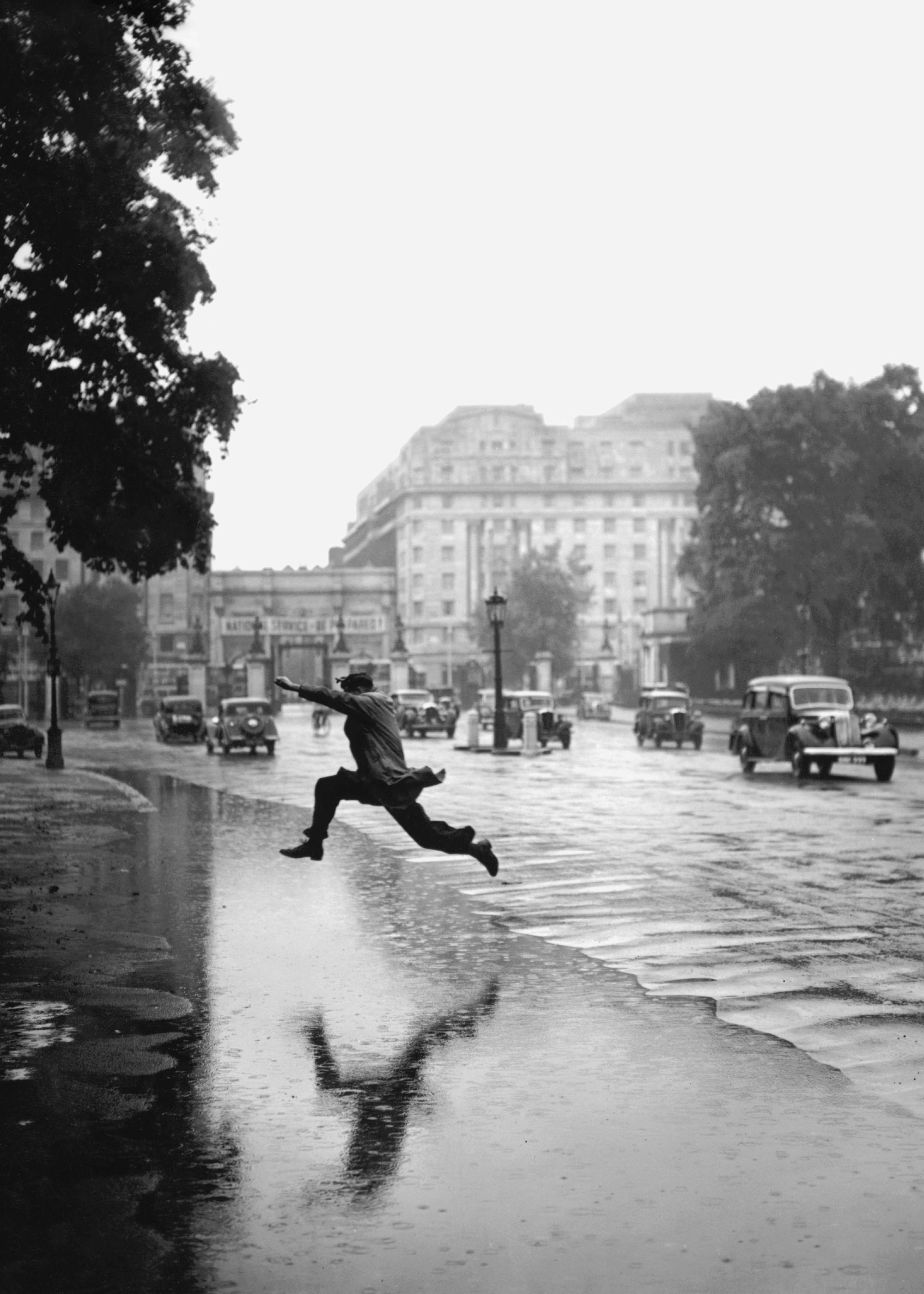 A black and white poster of a man leaping over a street puddle, his reflection visible, with old cars and buildings in the backg