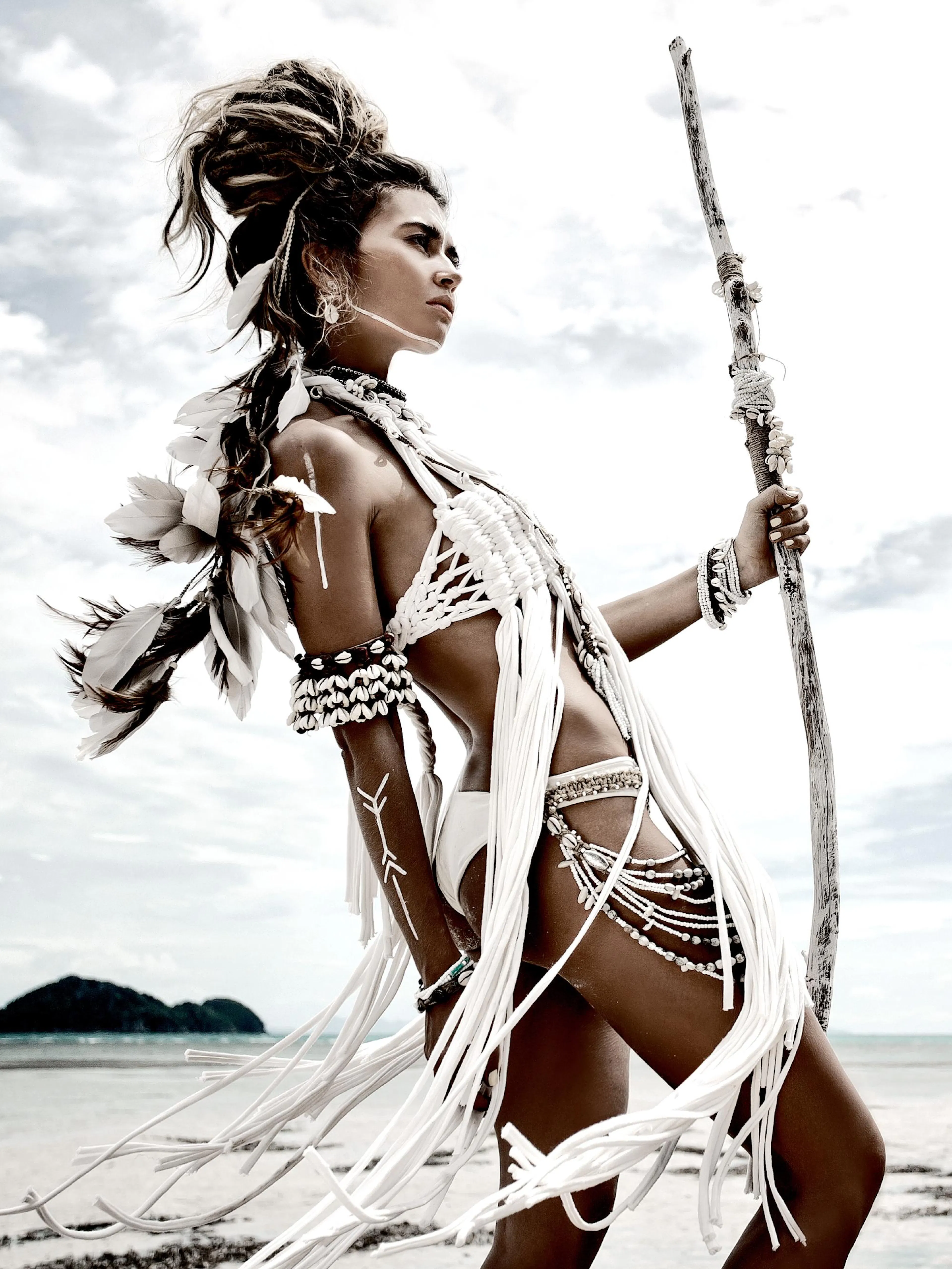 A poster featuring a woman with feathers and shells in her hair and clothing, holding a staff on a beach.