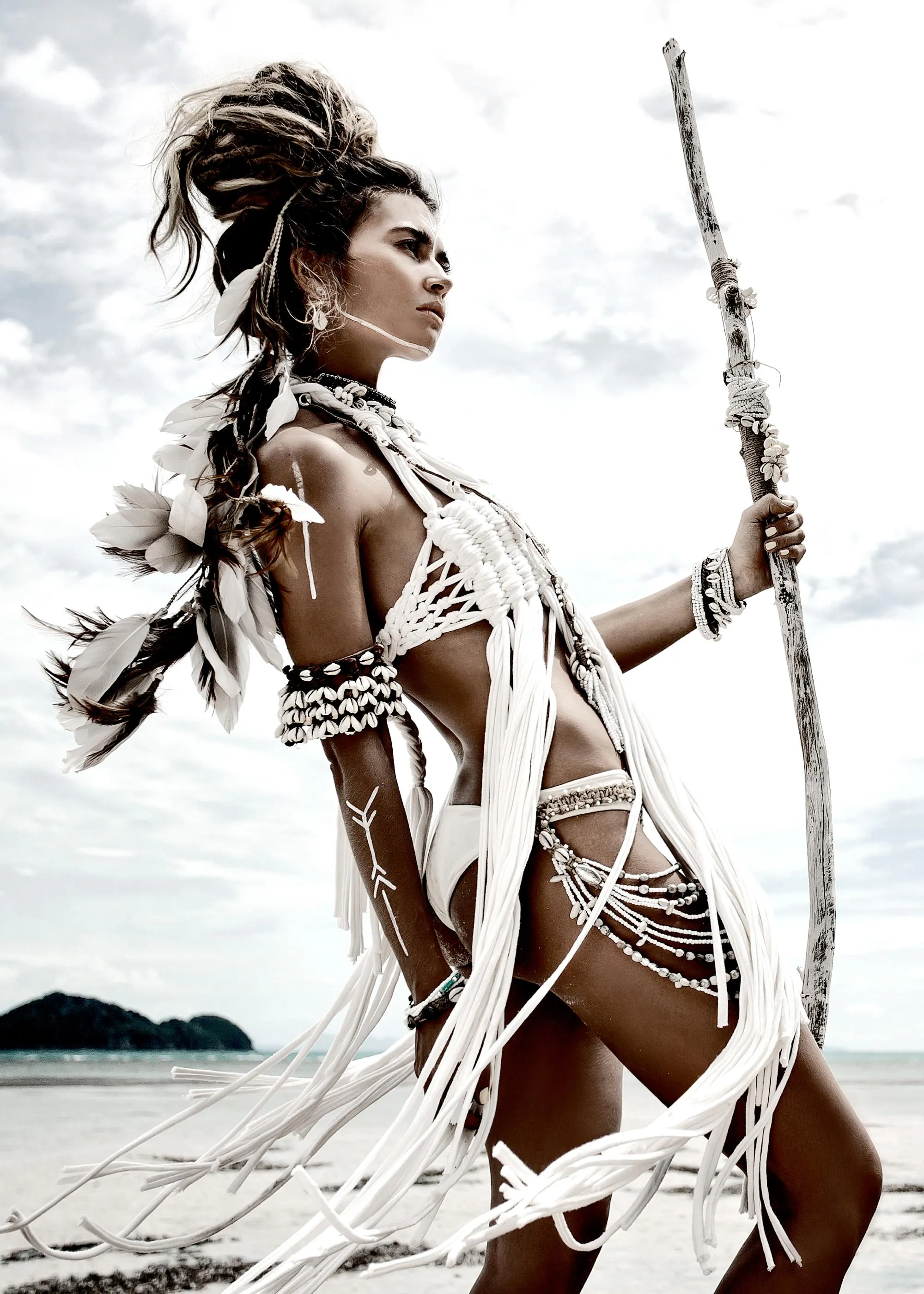 Poster dune femme bohème au maquillage tribal, vêtue de plumes et de coquillages, tenant un bâton sur la plage.