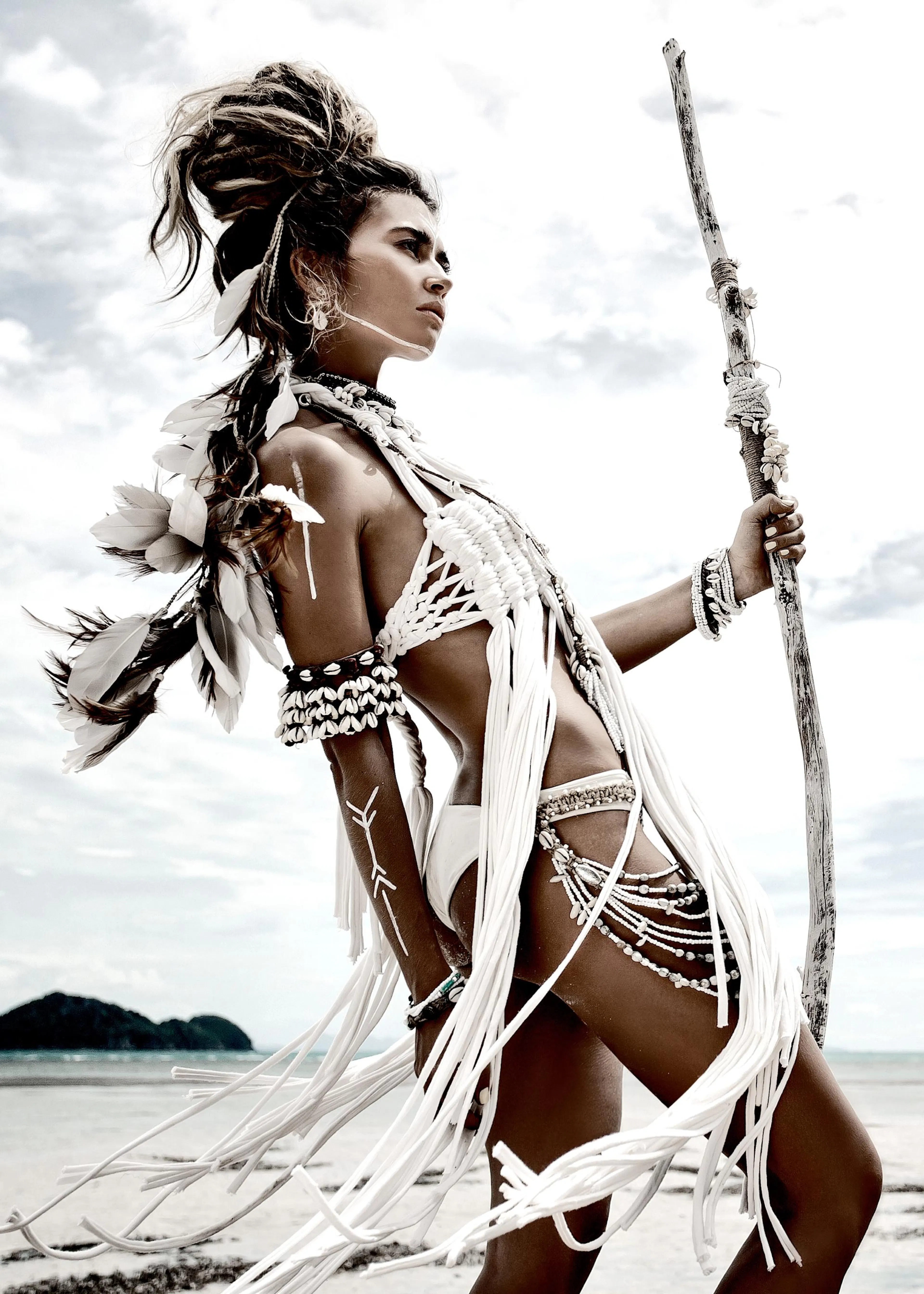 Un poster di una donna in abiti tribali bianchi con piume e conchiglie, che tiene un bastone, su una spiaggia.