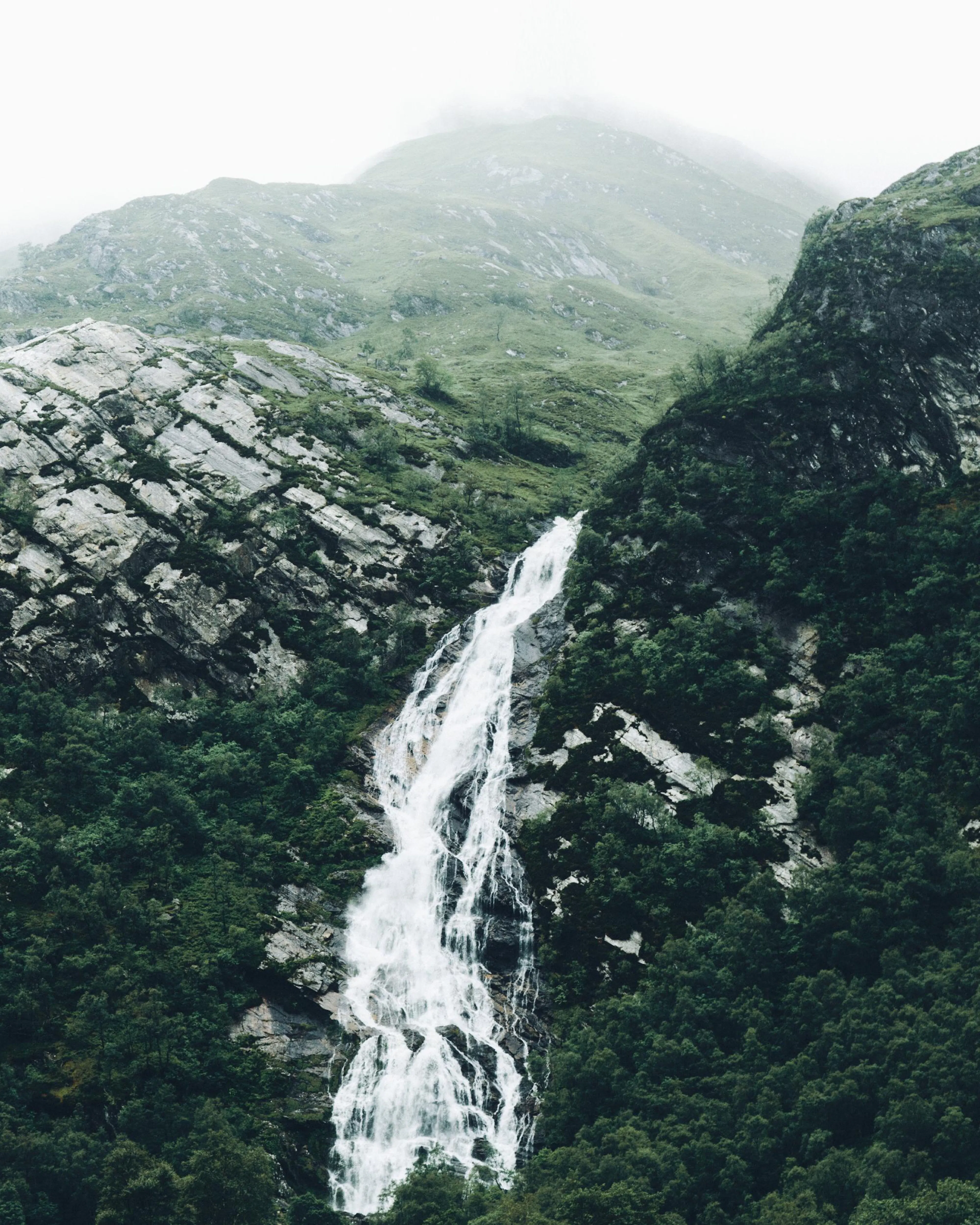 Poster con cascata che scende tra rocce grigie e vegetazione verde scuro in un paesaggio nebbioso di montagna.