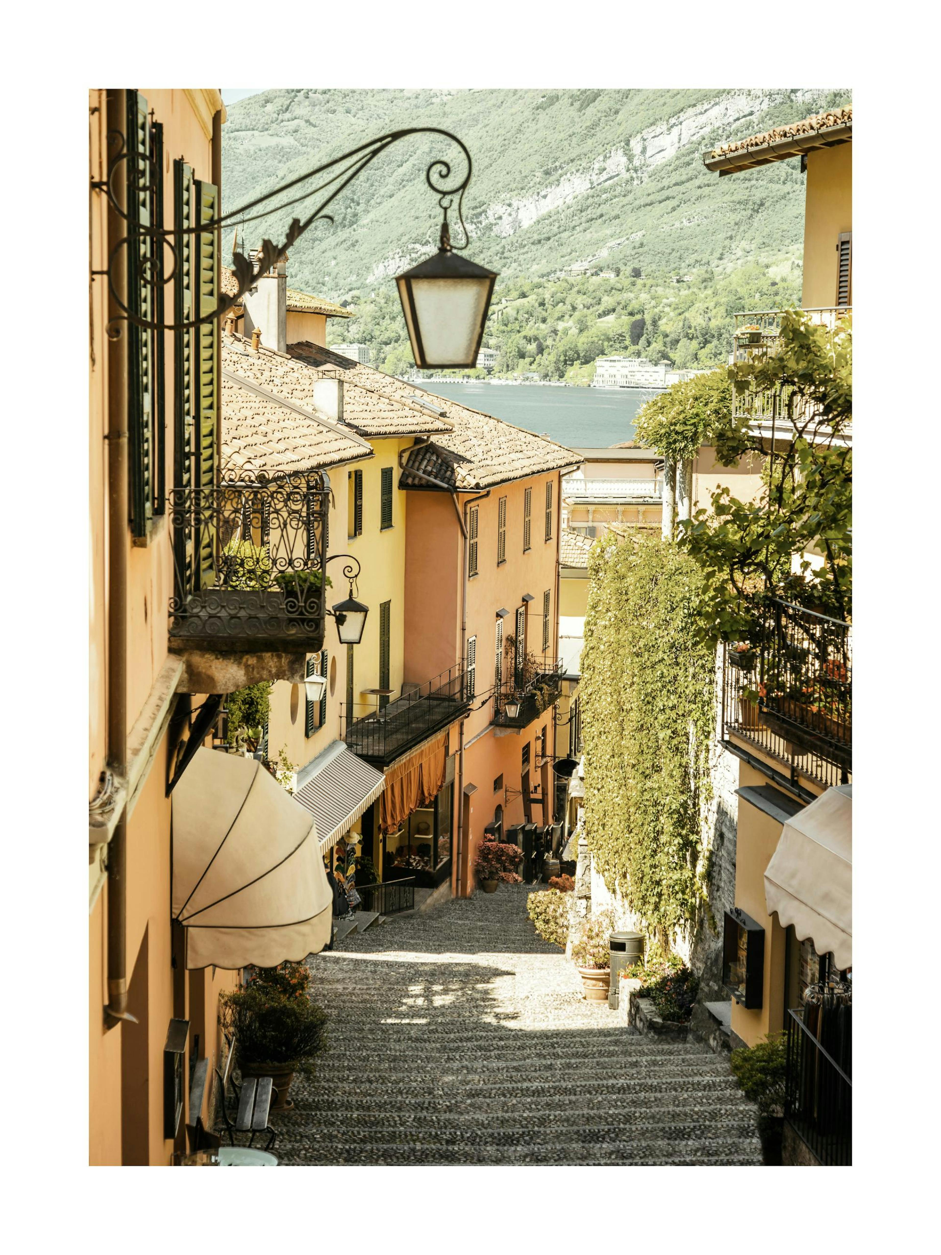 Póster de un callejón adoquinado con casas coloridas y un lago al fondo en un pueblo italiano.